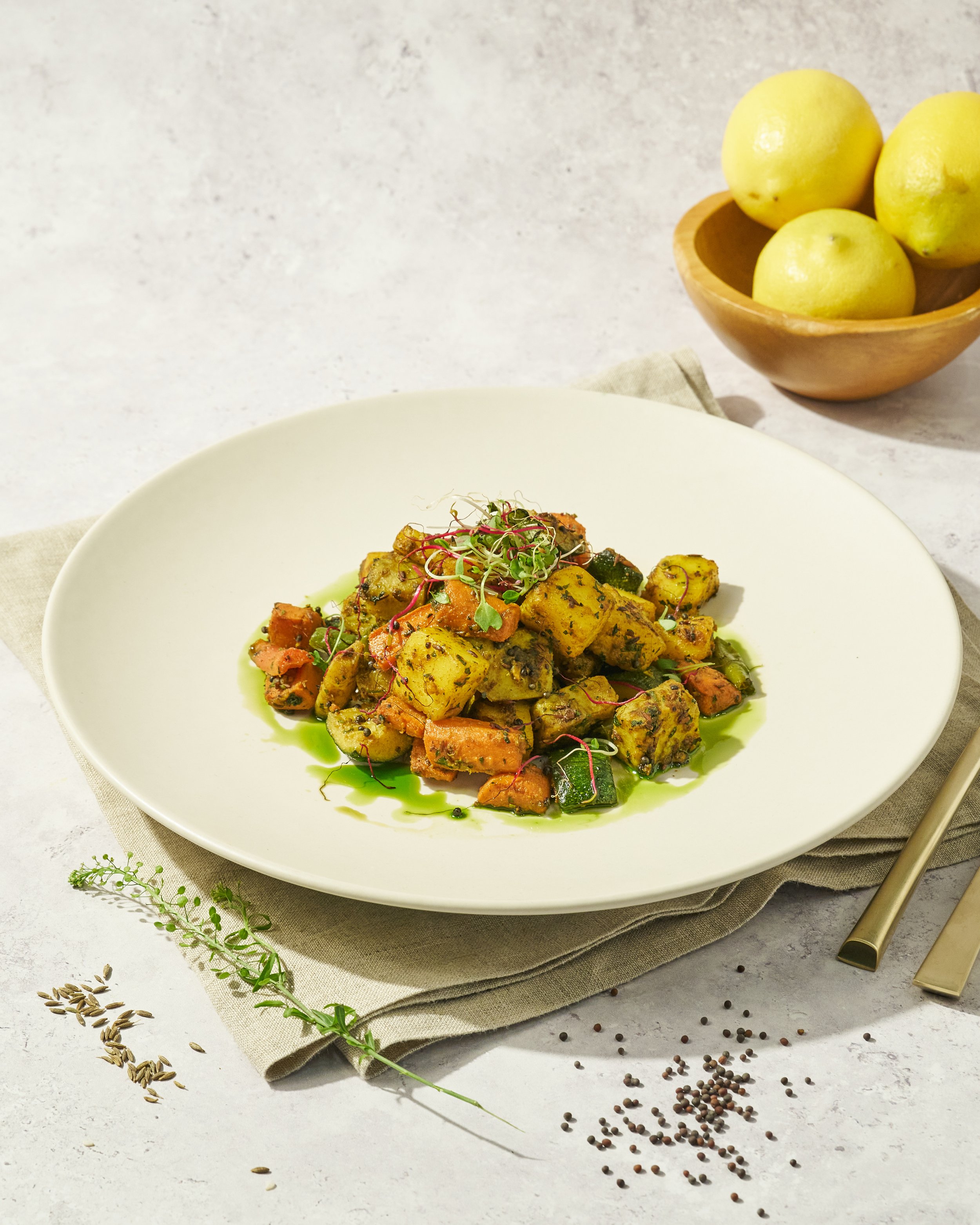 A white bowl of roasted vegetable salad with herbs on a light gray surface. In the background, three lemons in a wooden bowl. Nearby, scattered herbs, black pepper, and cumin seeds are arranged on the surface, with a gold fork partially visible on th
