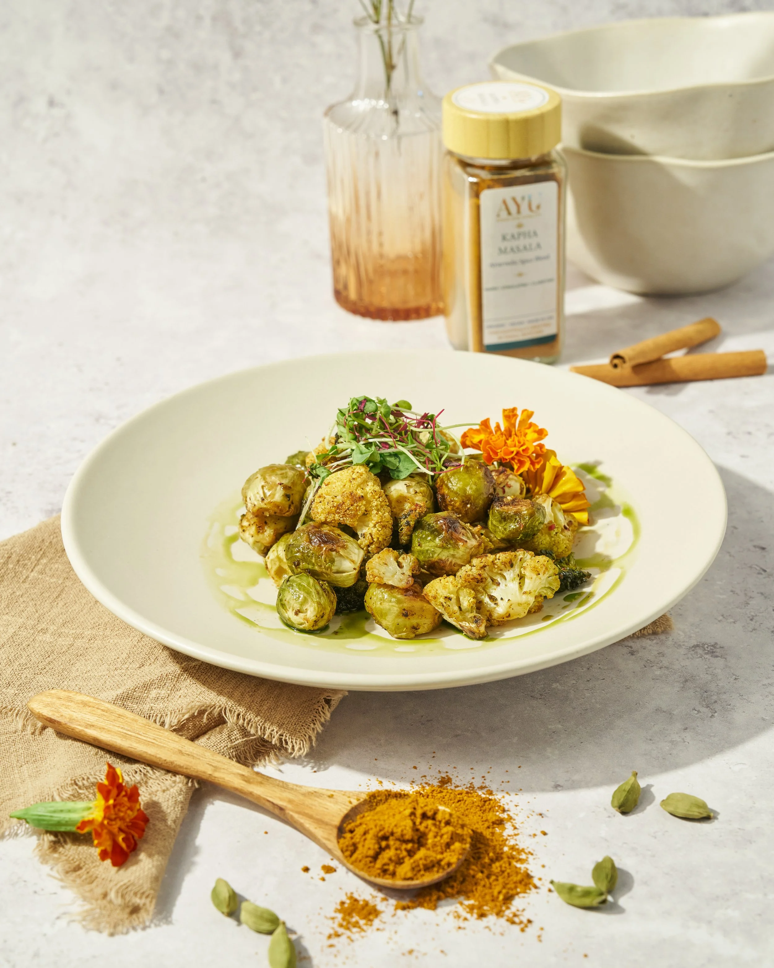 A plate of roasted Brussels sprouts garnished with microgreens and edible flowers, with a grated carrot on top, alongside a wooden spoon filled with turmeric powder, a decorative beige cloth, and scattered cardamom pods and turmeric on a white surfac