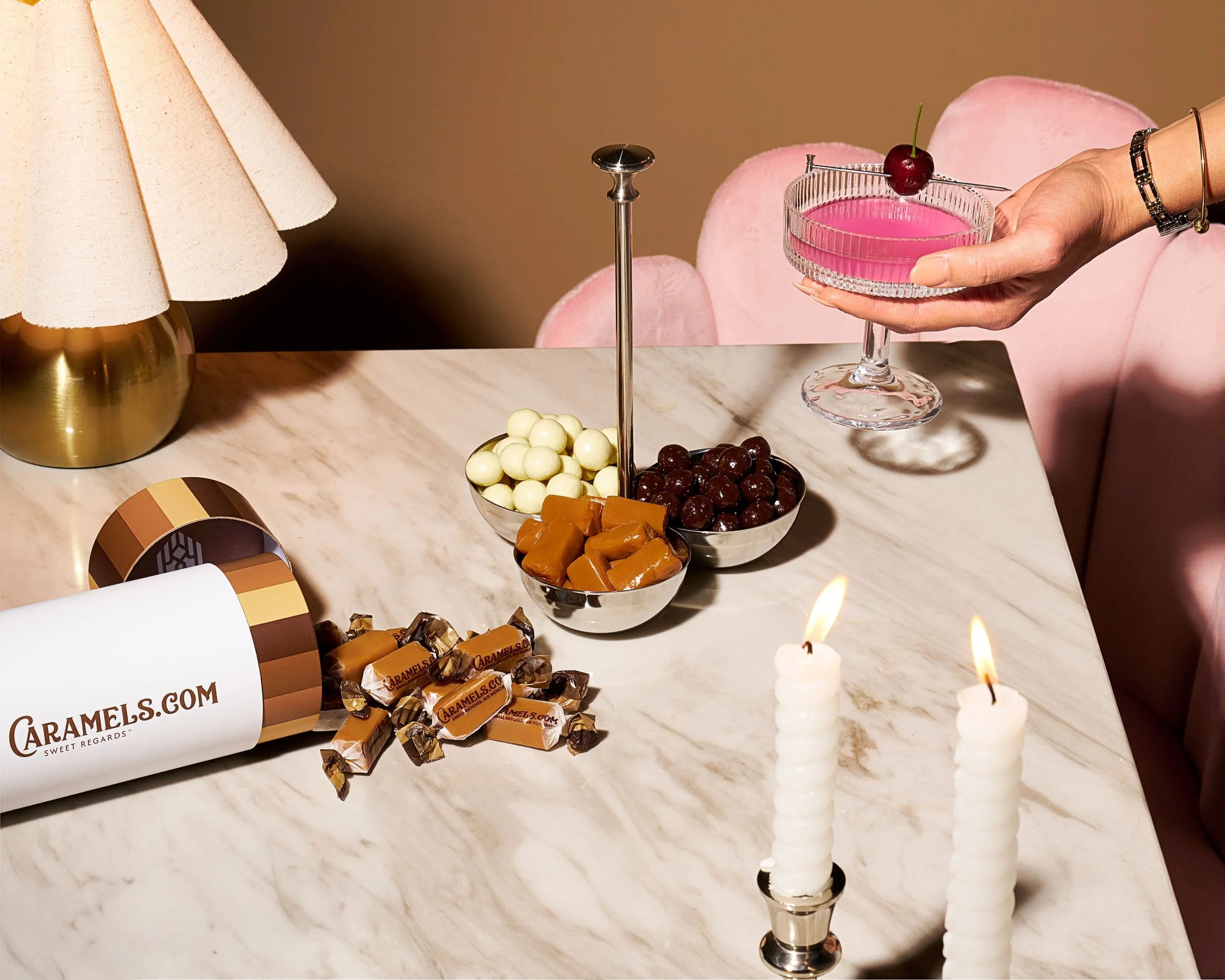 A photograph of a luxurious table setting featuring a hand holding a martini glass with a cherry garnish, white candles, assorted chocolates, and a white lamp. A hand in a pink sleeve is visible. The marble table has a "camels.com" chocolate box.