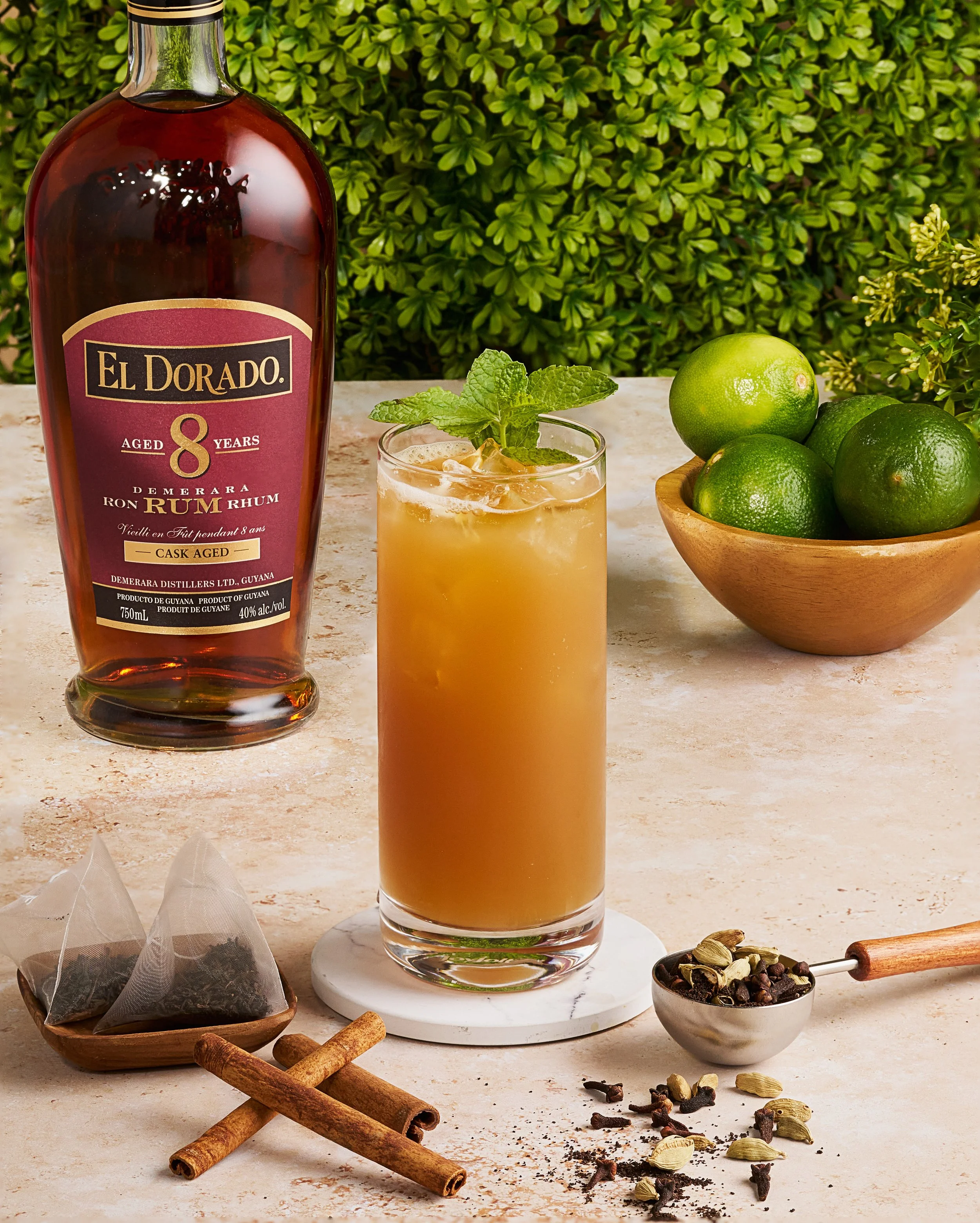 A photograph featuring a tall glass of iced tea garnished with mint, a bottle of El Dorado 8 Year Old Rum, and a bowl of green limes on a beige marble surface. The background includes lush green foliage. The setting evokes a tropical, refreshing ambi