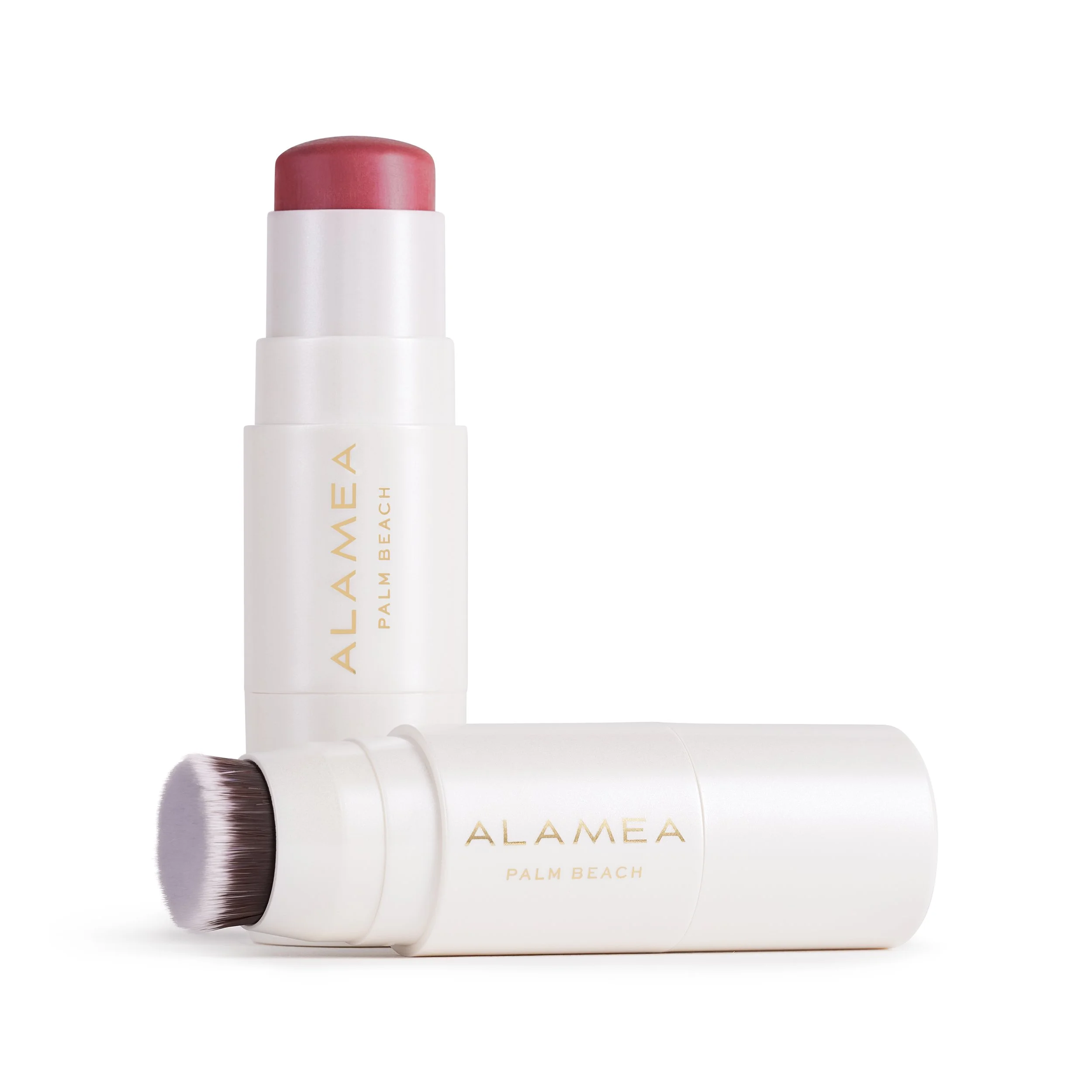 Two white cosmetic stick tubes from Alamea Palm Beach, one standing upright with a pink cream blush exposed, and the other lying horizontally with a built-in brush visible.
