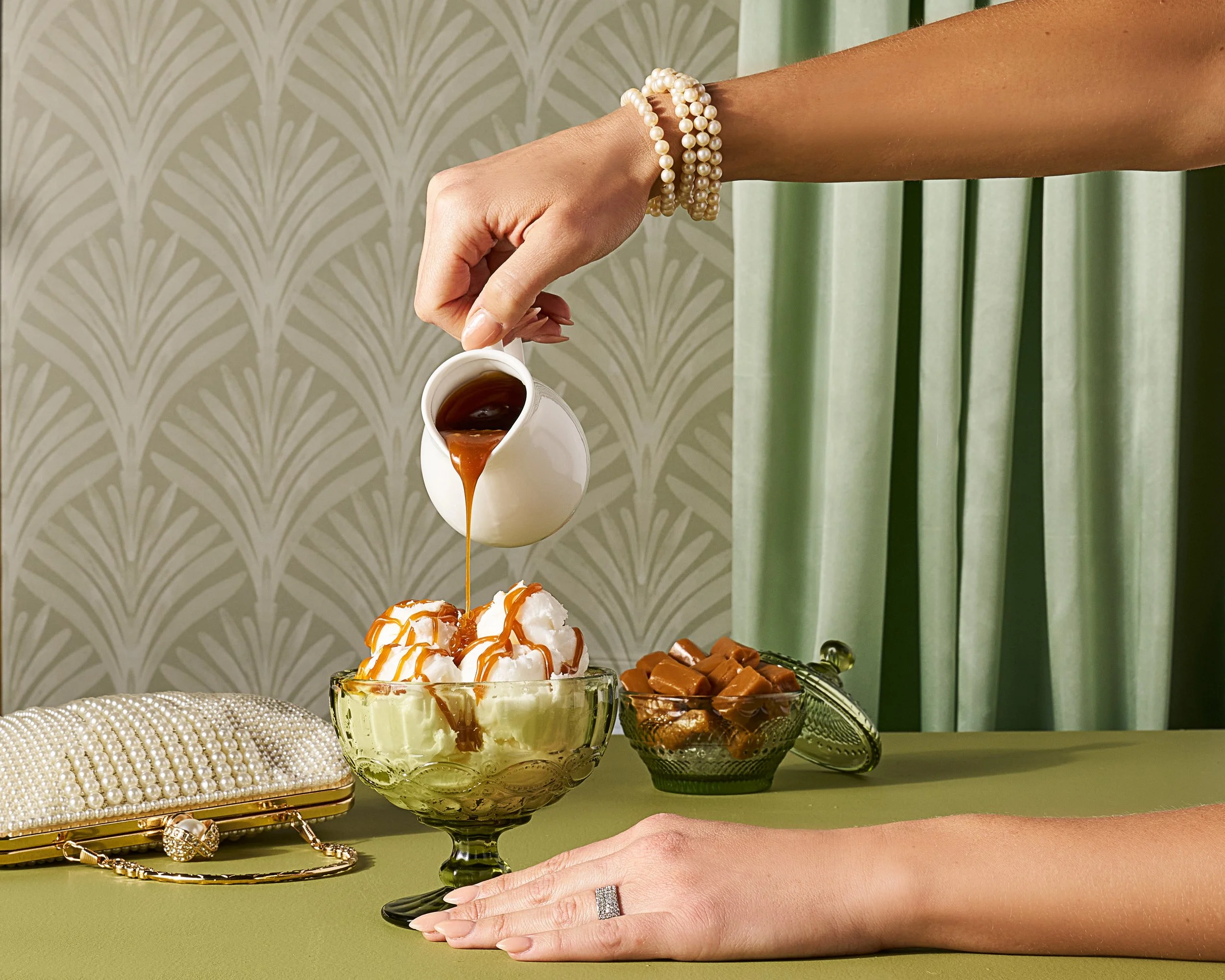 A elegant, warmly lit photograph of a person poruing caramel sauce on vanilla ice cream in. a bowl, with green accents and a art deco inspired background.