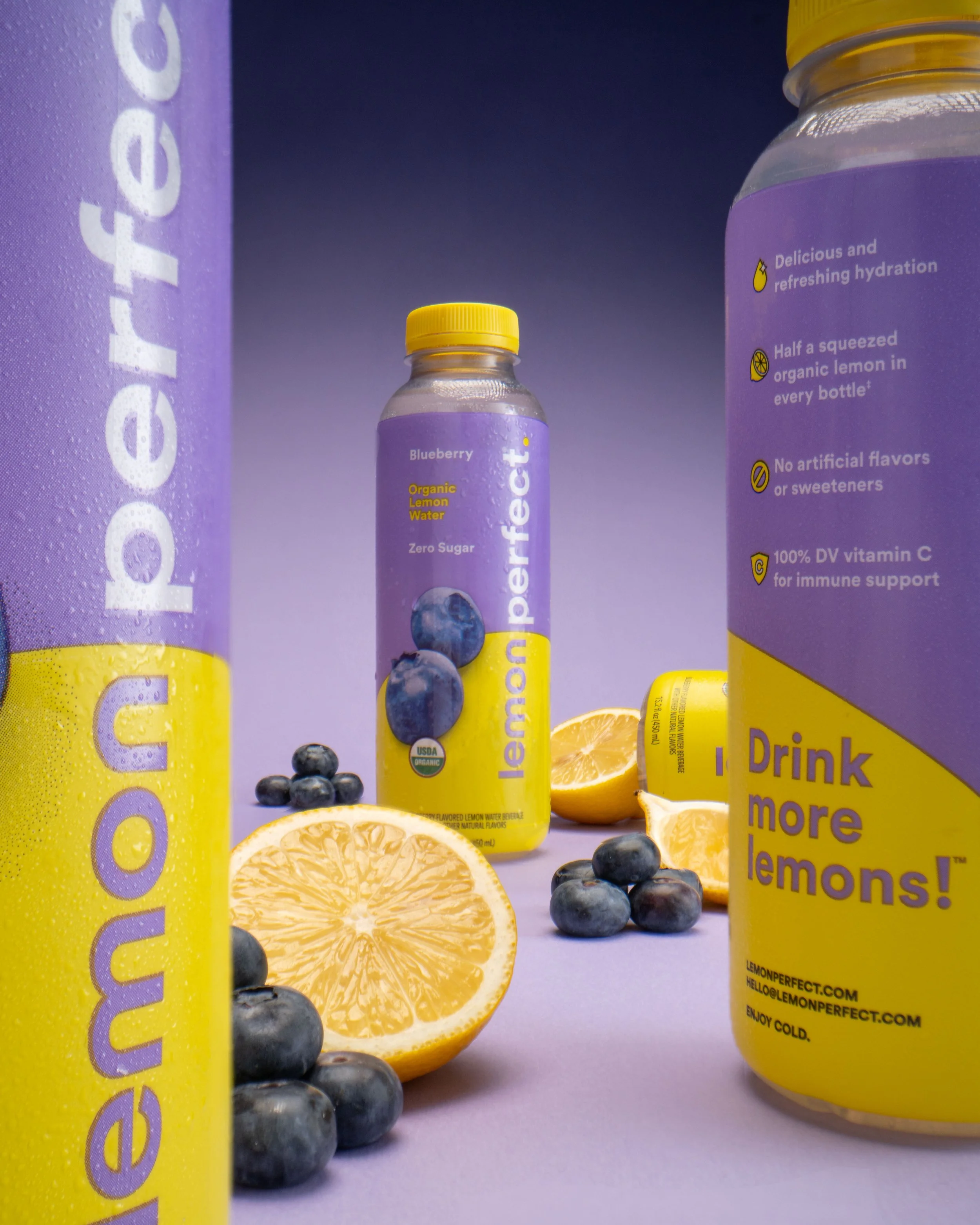 Blueberry flavored Lemon Perfect beverage, two bottles in the forefront allowing the eye to go directly to the bottle in the distant middle on a purple/lavender background with blueberries and oranges sprinkled throughout.