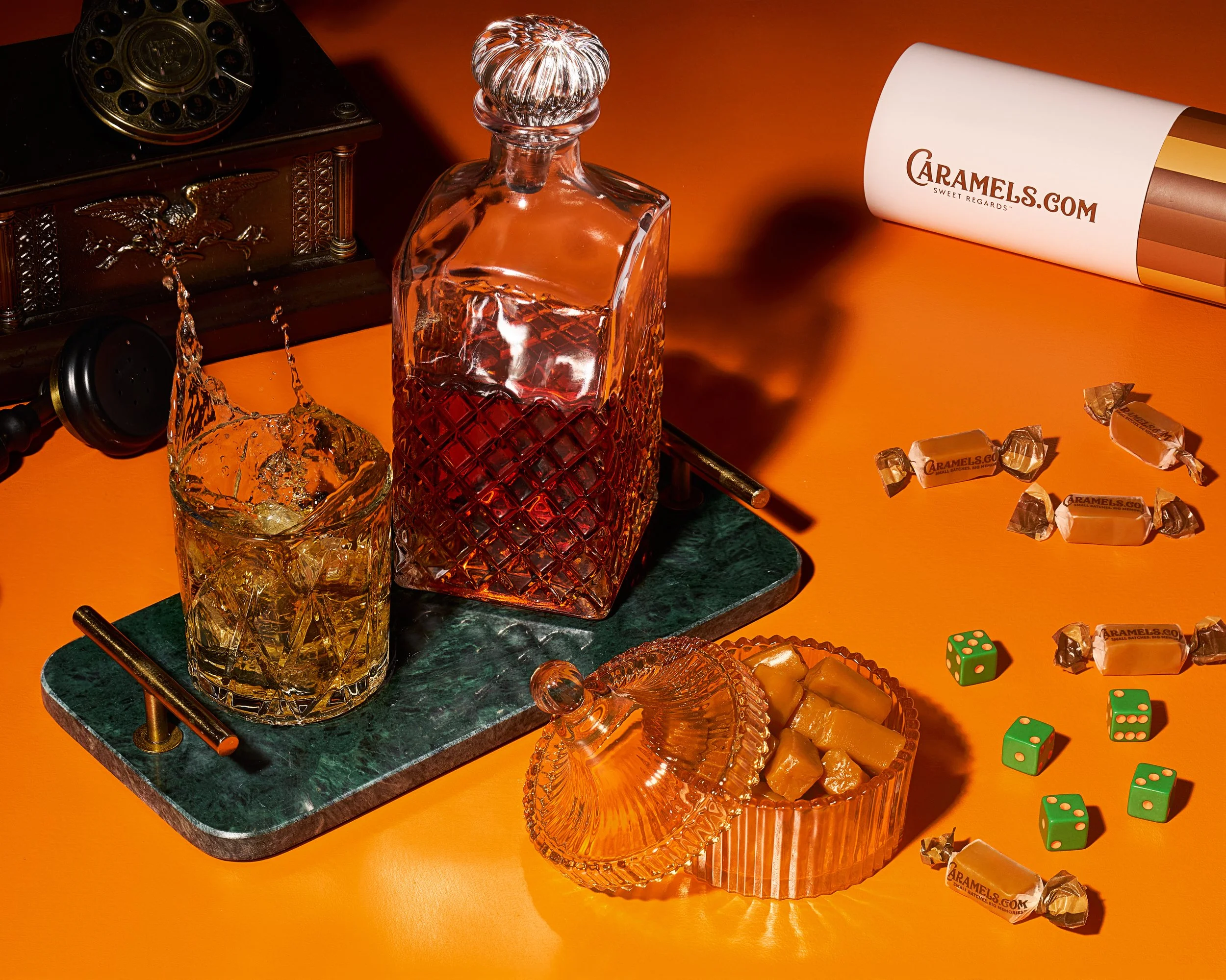 A photograph featuring a vintage scene with a dark wooden rotary phone, a green marble tray holding a crystal decanter of amber liquid and a glass filled with ice, scattered dice, and a clear glass candy dish filled with brown candies.