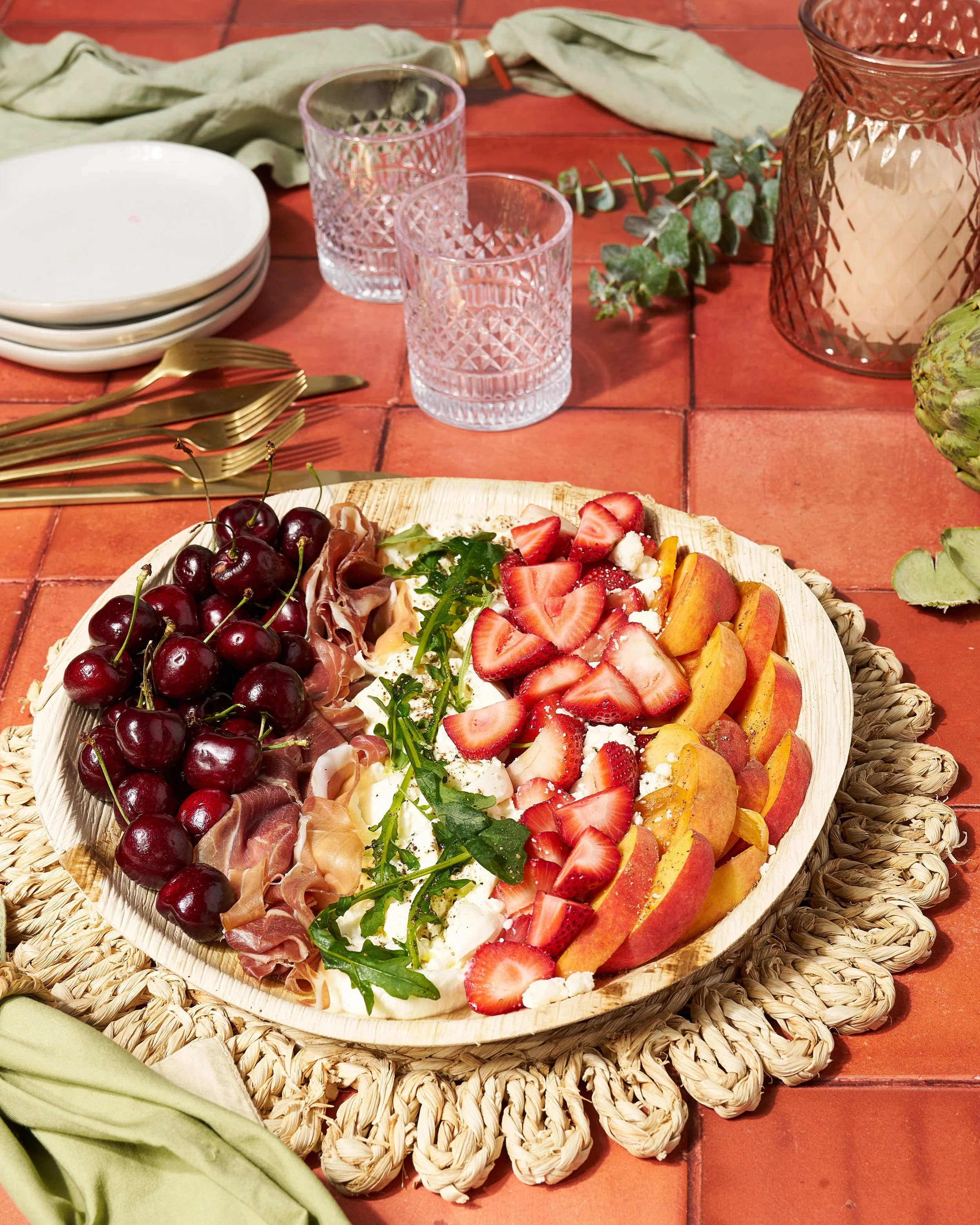Photograph of a rustic wooden platter with a colorful fruit salad of red cherries, sliced peaches, strawberries, and kiwi. The platter sits on a textured, beige woven placemat on an orange-tiled table. Surrounding the platter are neatly folded green