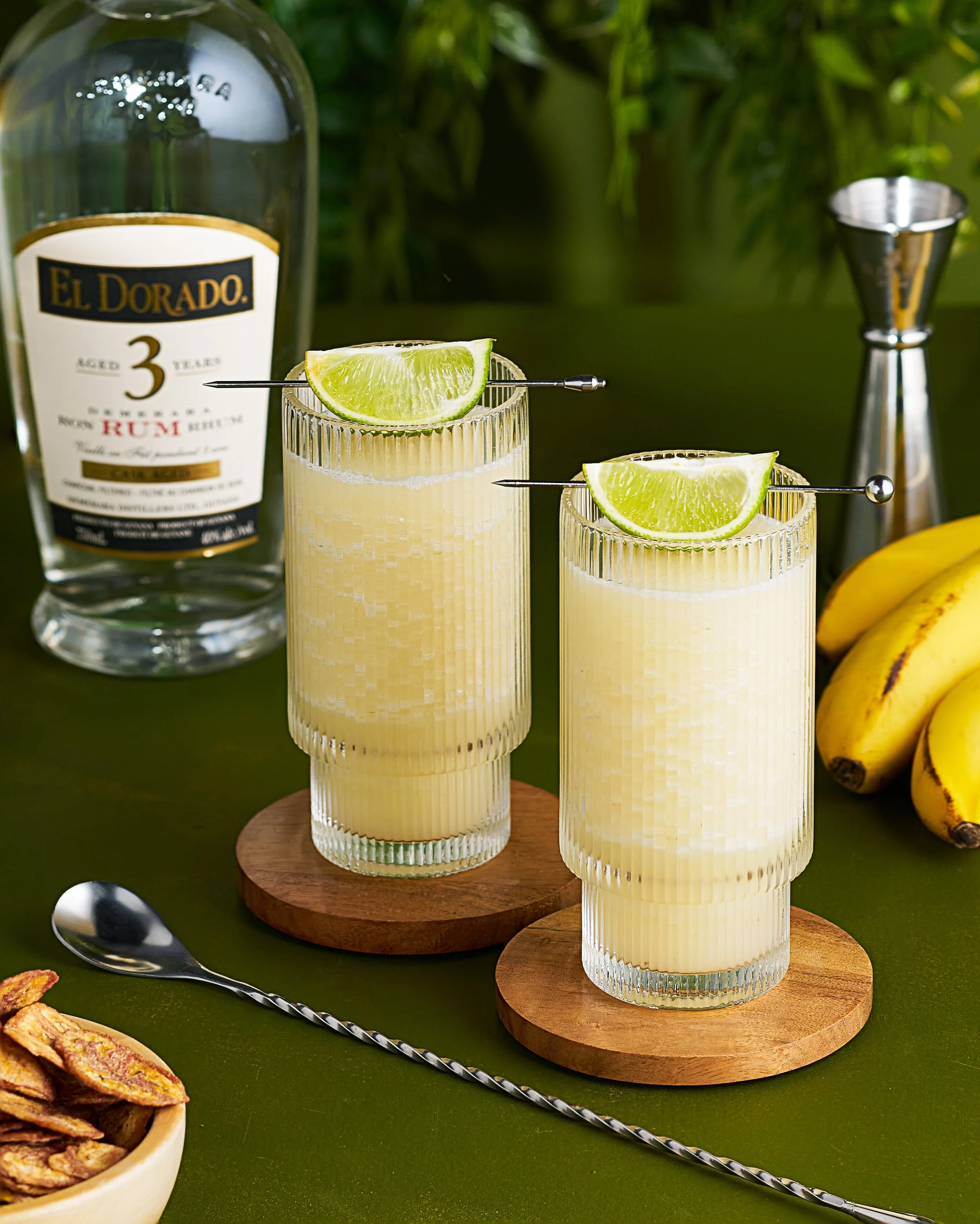 Photograph of two clear, ribbed glasses filled with pale yellow, frothy rum punch, garnished with lime wedges and straws. Background includes a bottle of El Dorado 3 Year Old Rum, bananas, a silver shaker, and a wooden bowl of potato chips on a green