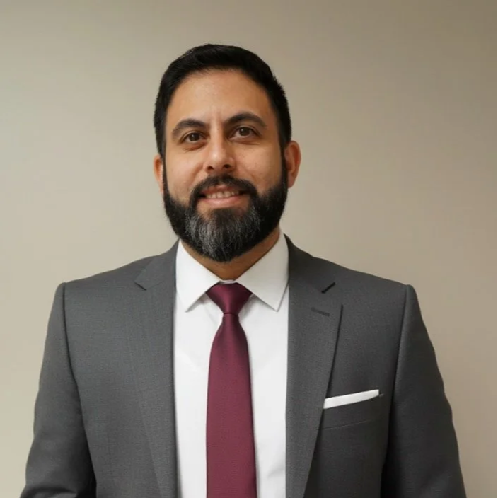 Portrait of a man in a gray suit with a white shirt, burgundy tie, and pocket square, smiling against a plain background.