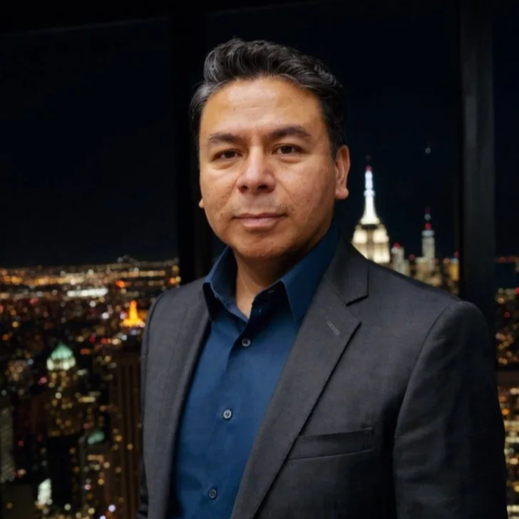 A man in a suit standing in front of a city skyline at night with illuminated buildings and a prominent tower in the background.