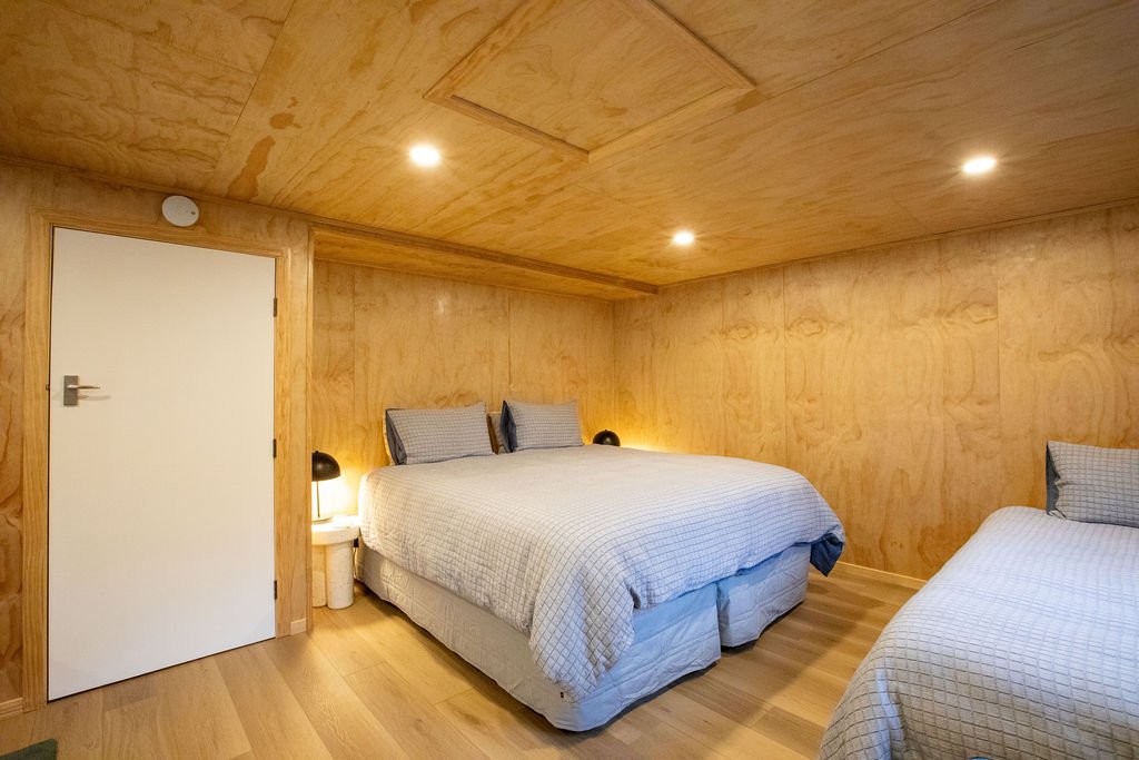 Bedroom with wooden interior, two beds with checkered bedding and pillows, small nightstands with lamps, ceiling with recessed lighting.