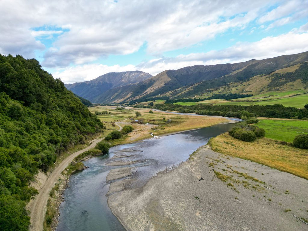 A wide river flowing through a green valley surrounded by rolling hills and mountains under a partly cloudy sky.