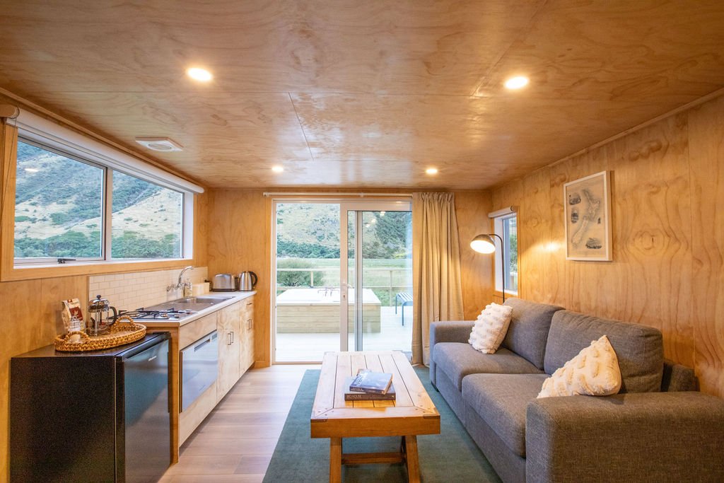 Cozy living room with wood-paneled walls and ceiling, featuring a gray couch with white pillows, a wooden coffee table, a small kitchen area with a fridge, countertop, and appliances, and large windows and sliding glass door leading to a balcony with