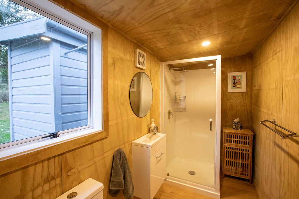 Small bathroom with wooden walls, a window, a shower with a glass door, a mirror above a white sink, a small cabinet, and a towel rack.