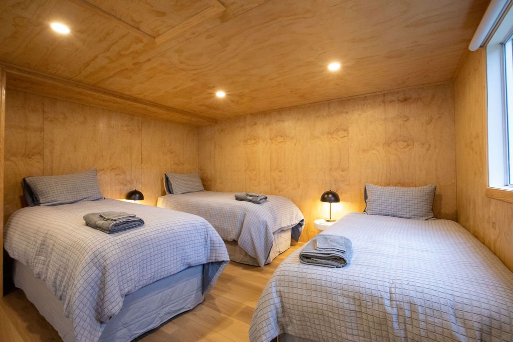 Three beds with gray checkered bedding and pillows, each with folded towels on top, in a wooden-paneled guest room with small bedside lamps and a window.