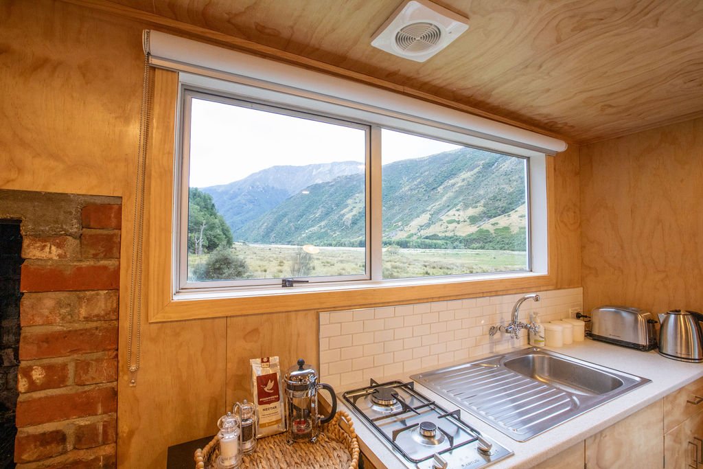 Kitchen with a window view of mountains and greenery, wooden walls and ceiling, countertop with stainless steel sink, gas stove, toaster, electric kettle, and various kitchen items.