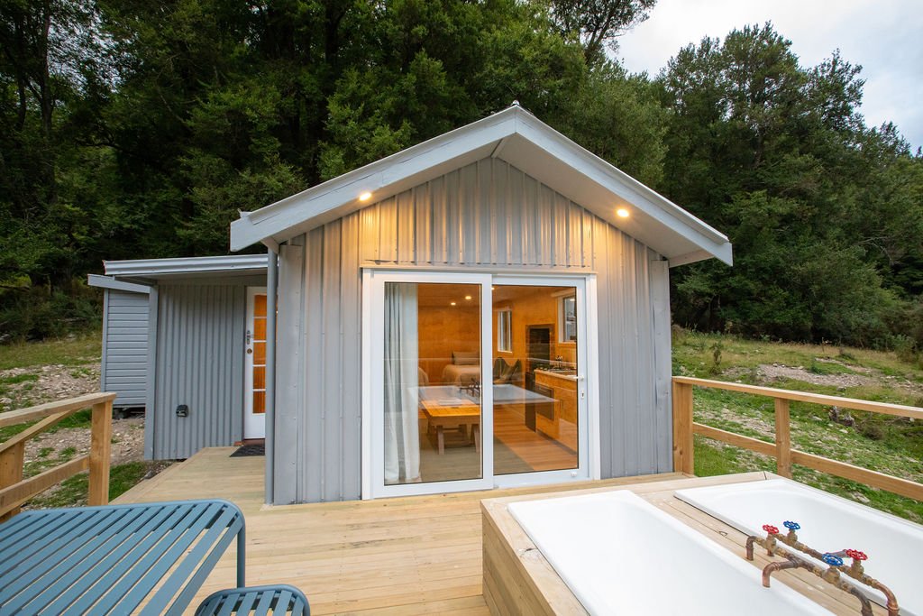 Exterior view of a small house with a deck, sliding glass door, and an outdoor hot tub, set in a wooded area.