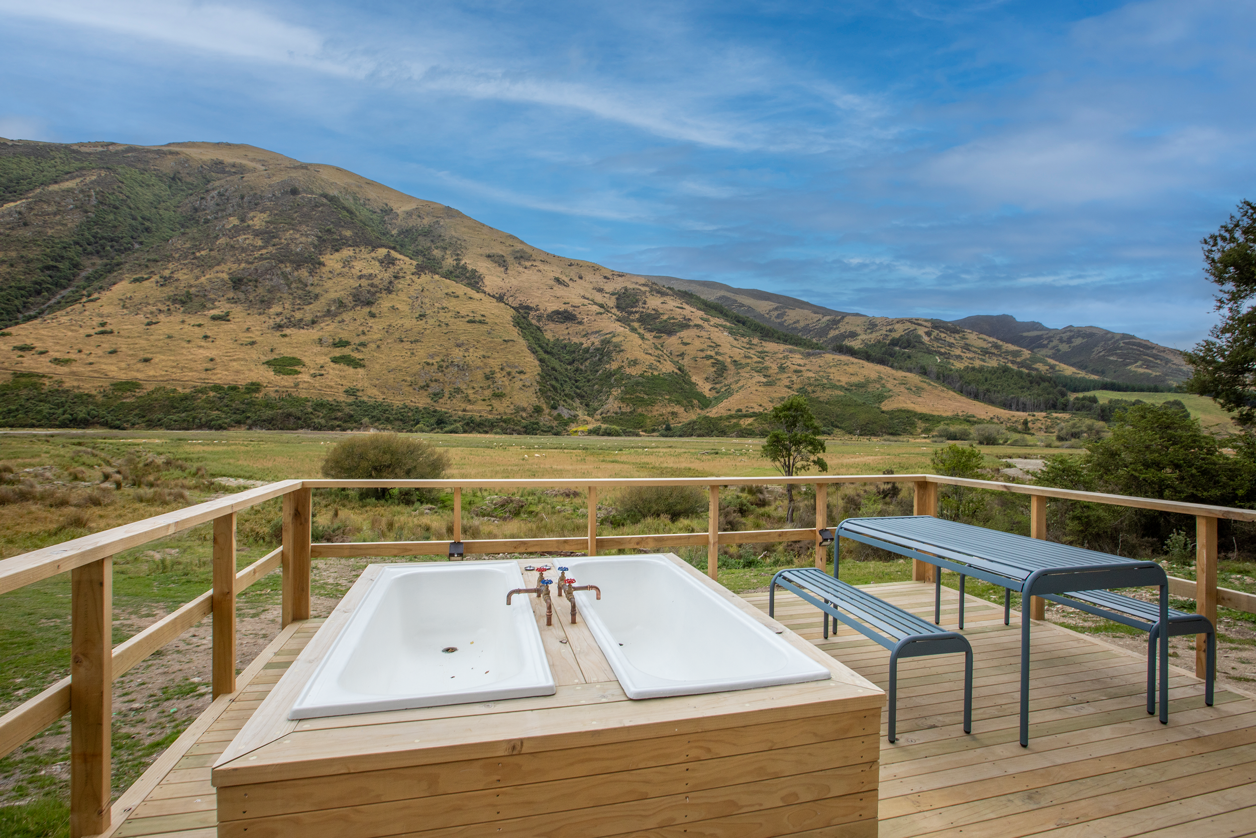 Wooden deck with a hot tub and a blue bench overlooking green fields and mountains under a partly cloudy sky.