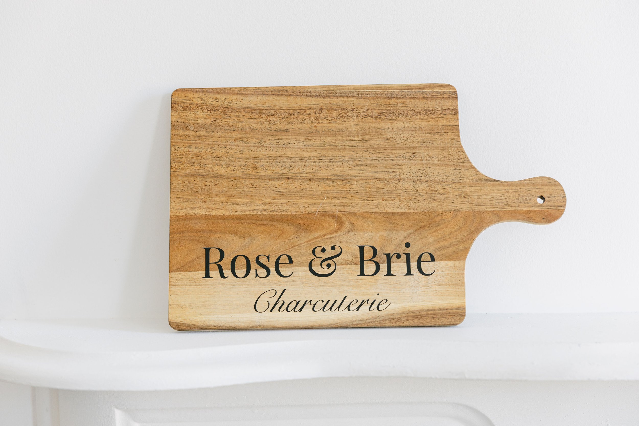 Wooden cutting board with the text 'Rose & Brie Charcuterie' printed on it, resting against a white wall.