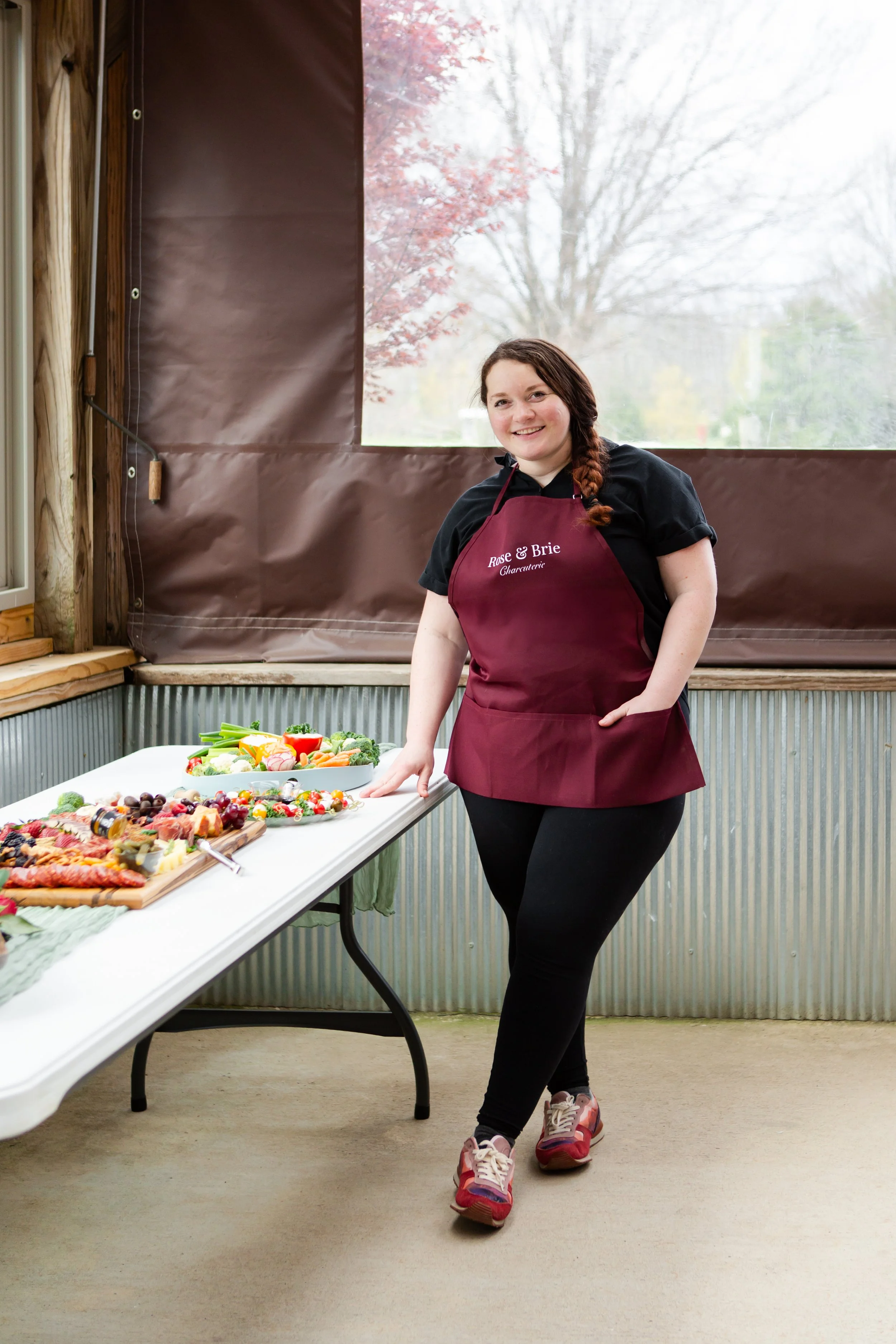 A woman wearing a maroon apron and black shirt standing next to a table with various food items, including fruits and vegetables, in a room with wood and metal walls and a large window showing trees outside.