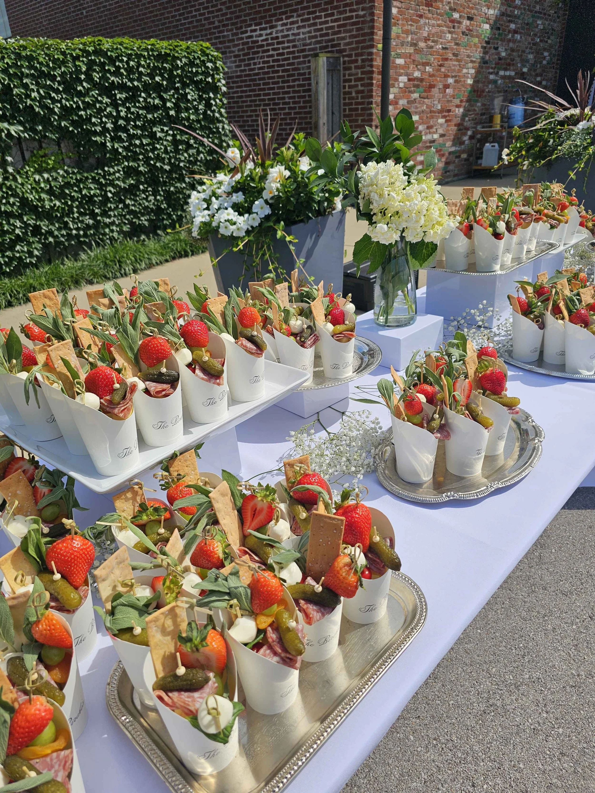 A table with assorted desserts in white cups and small bowls, garnished with strawberries and green leaves, set outdoors with a brick wall and greenery in the background.