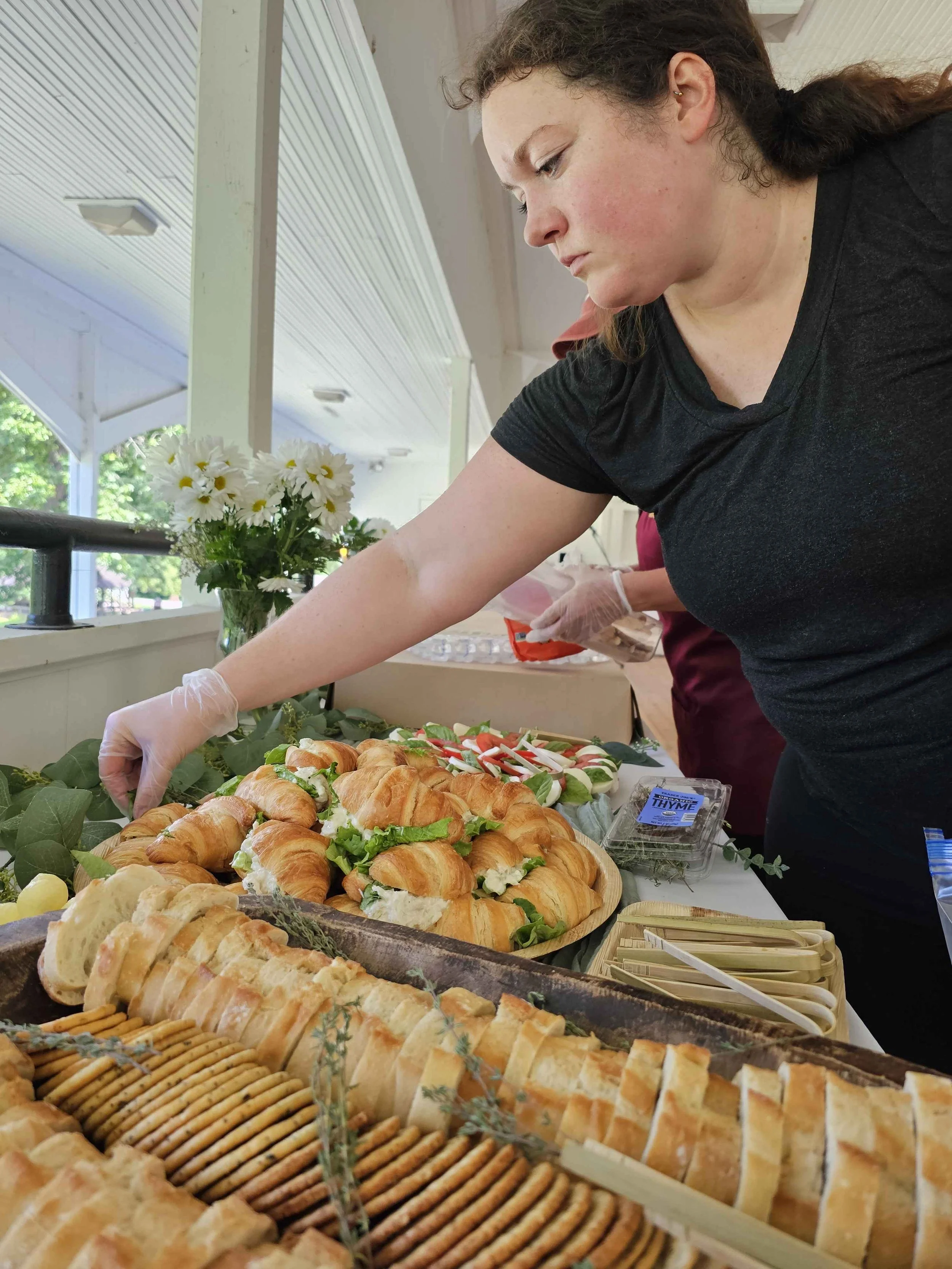 A woman in black shirt and gloves reaches for baked goods and fresh vegetables on a table at a buffet-style food display inside a bright, airy building with windows and trees outside.