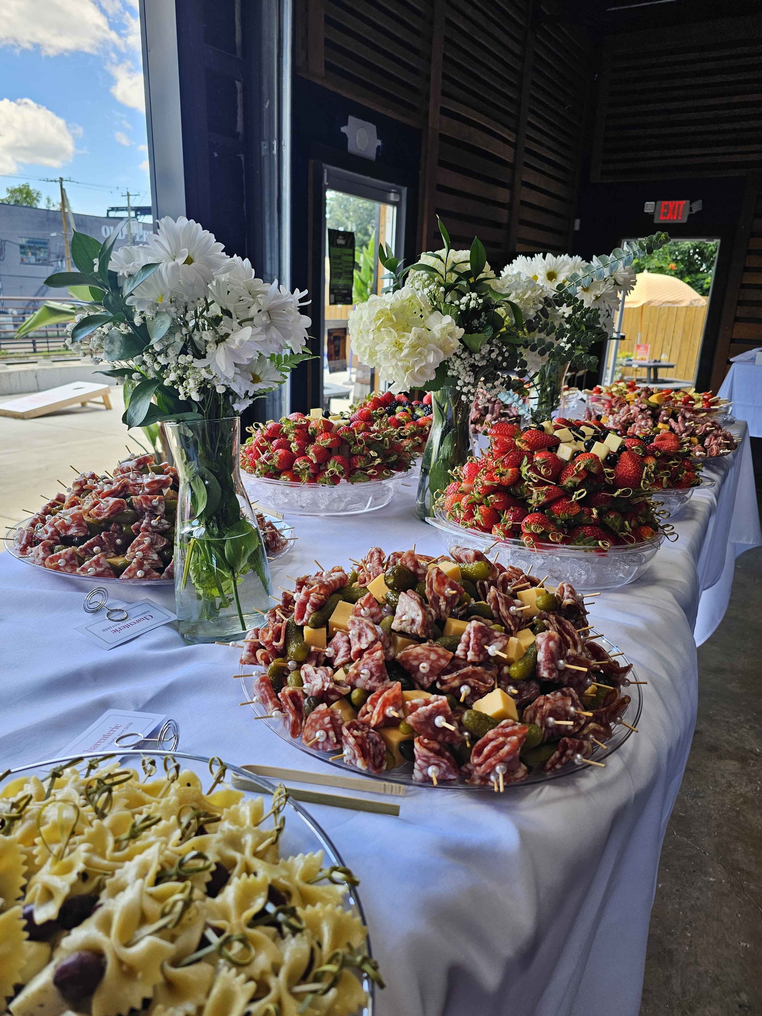A table at a party or event with three glass vases holding white flowers and greenery, and several platters of snacks including skewers of fruit, cheese, and meats, set up inside a venue with open doors to the outside.