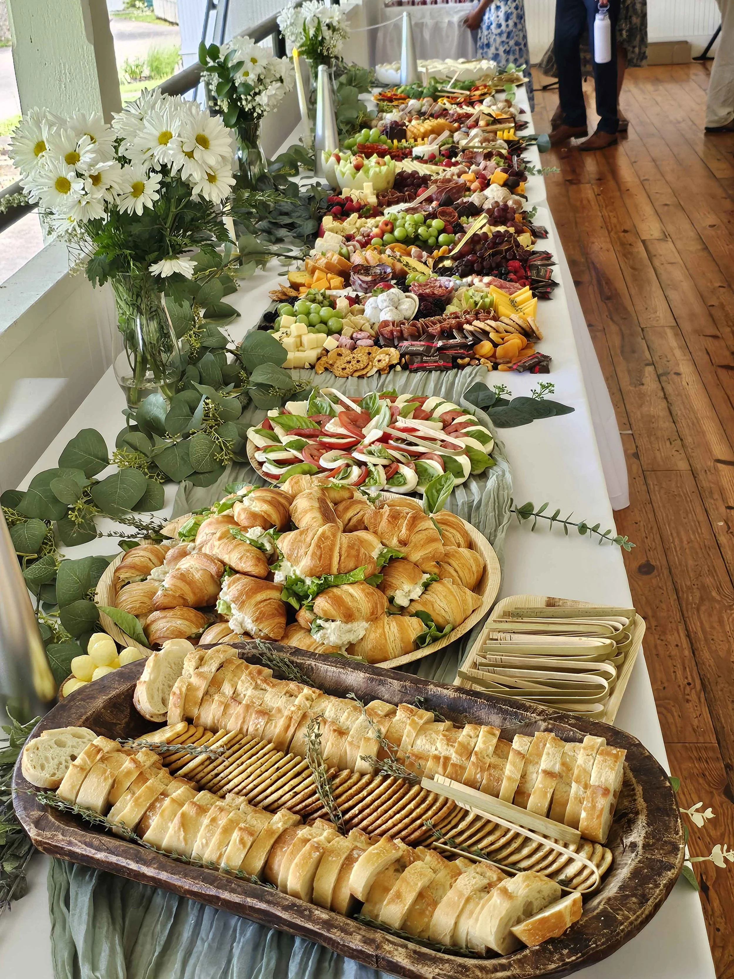 A buffet table with an assortment of cheeses, fruits, and bread, decorated with white flowers and green foliage at a daytime event.