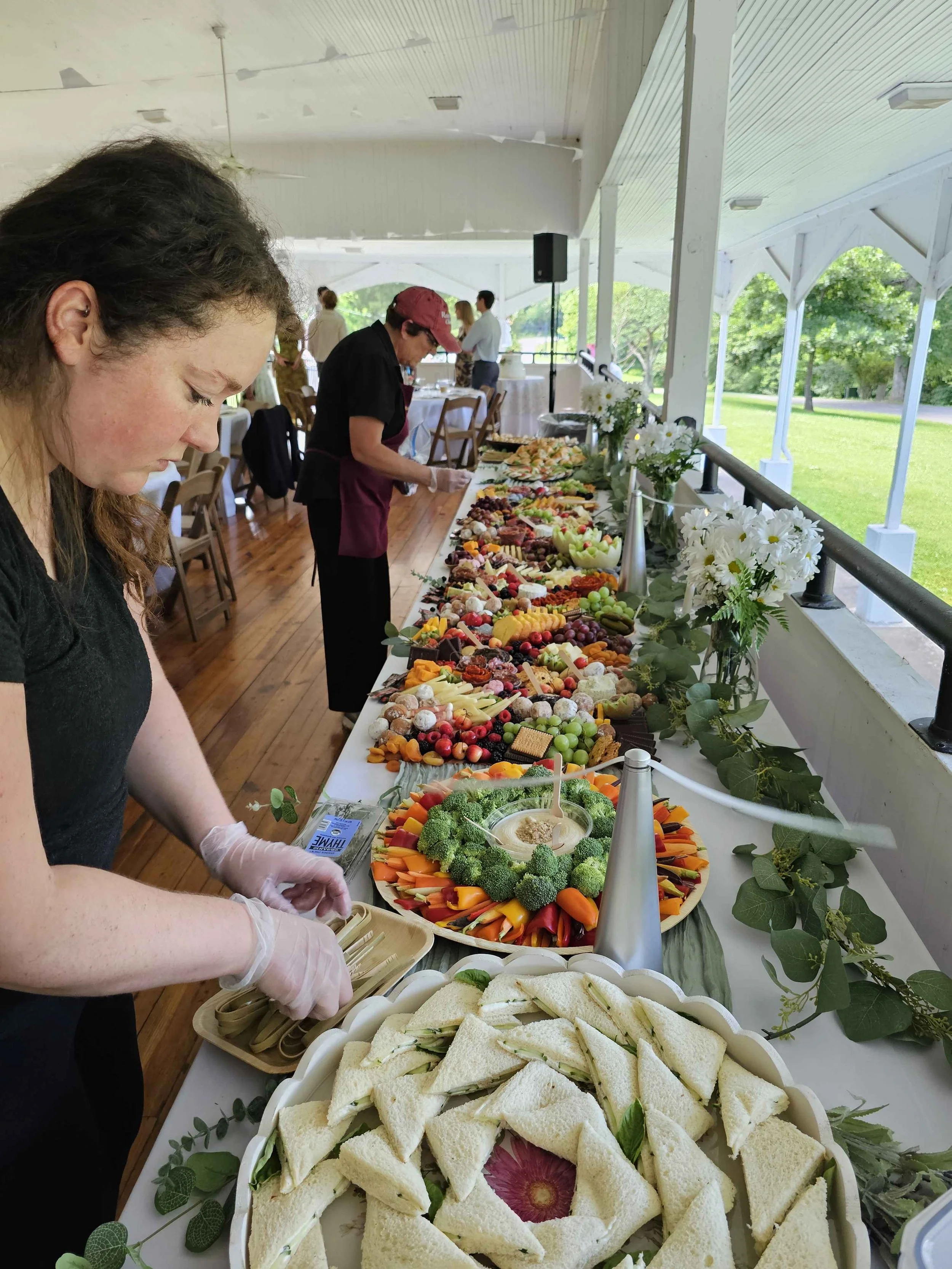 A woman preparing sandwiches at a buffet table with various fruits, vegetables, and sandwiches, in a covered outdoor venue with greenery visible outside.