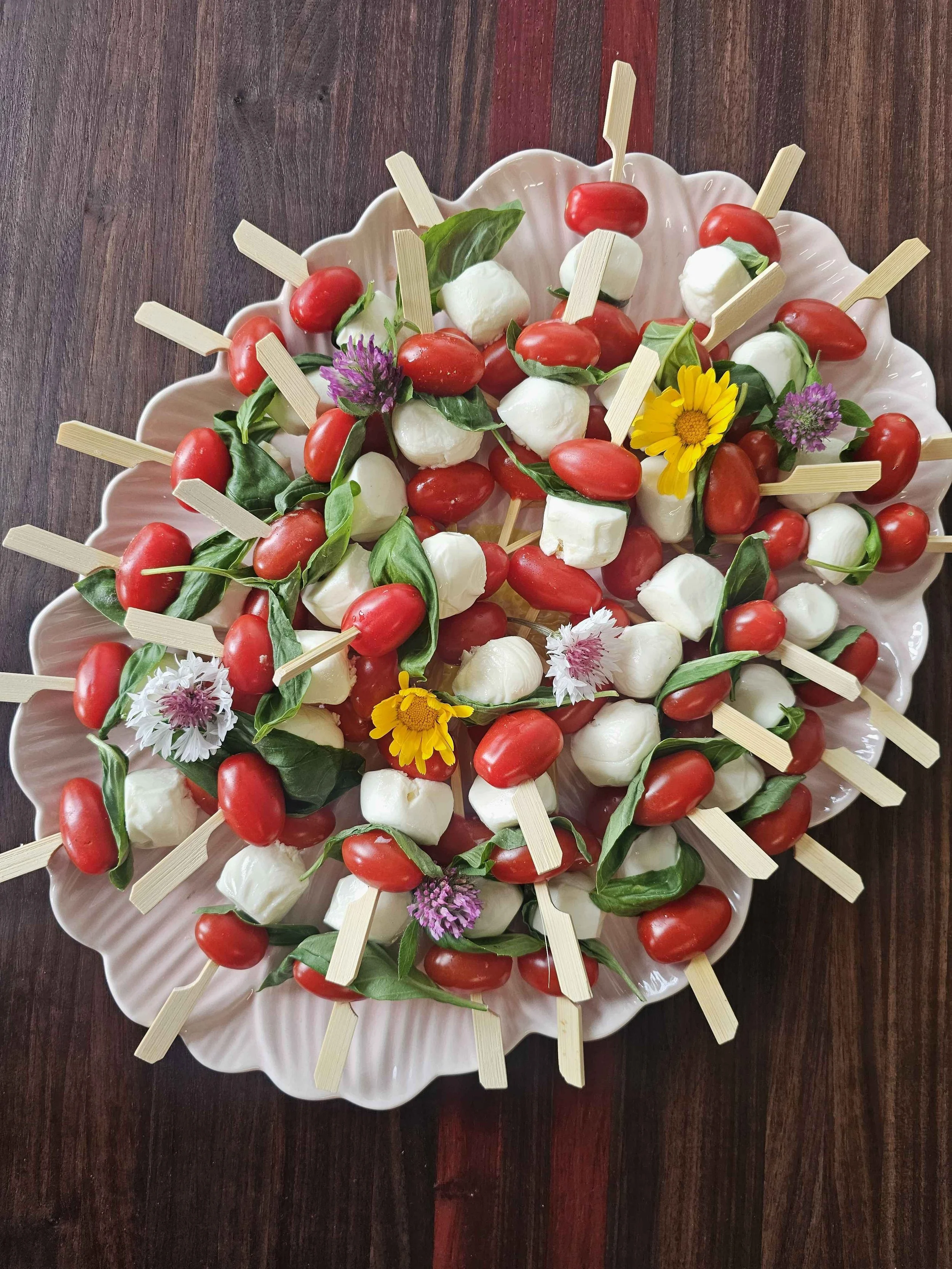 Plate of caprese skewers with cherry tomatoes, mozzarella balls, basil leaves, and edible flowers on a dark wooden table.