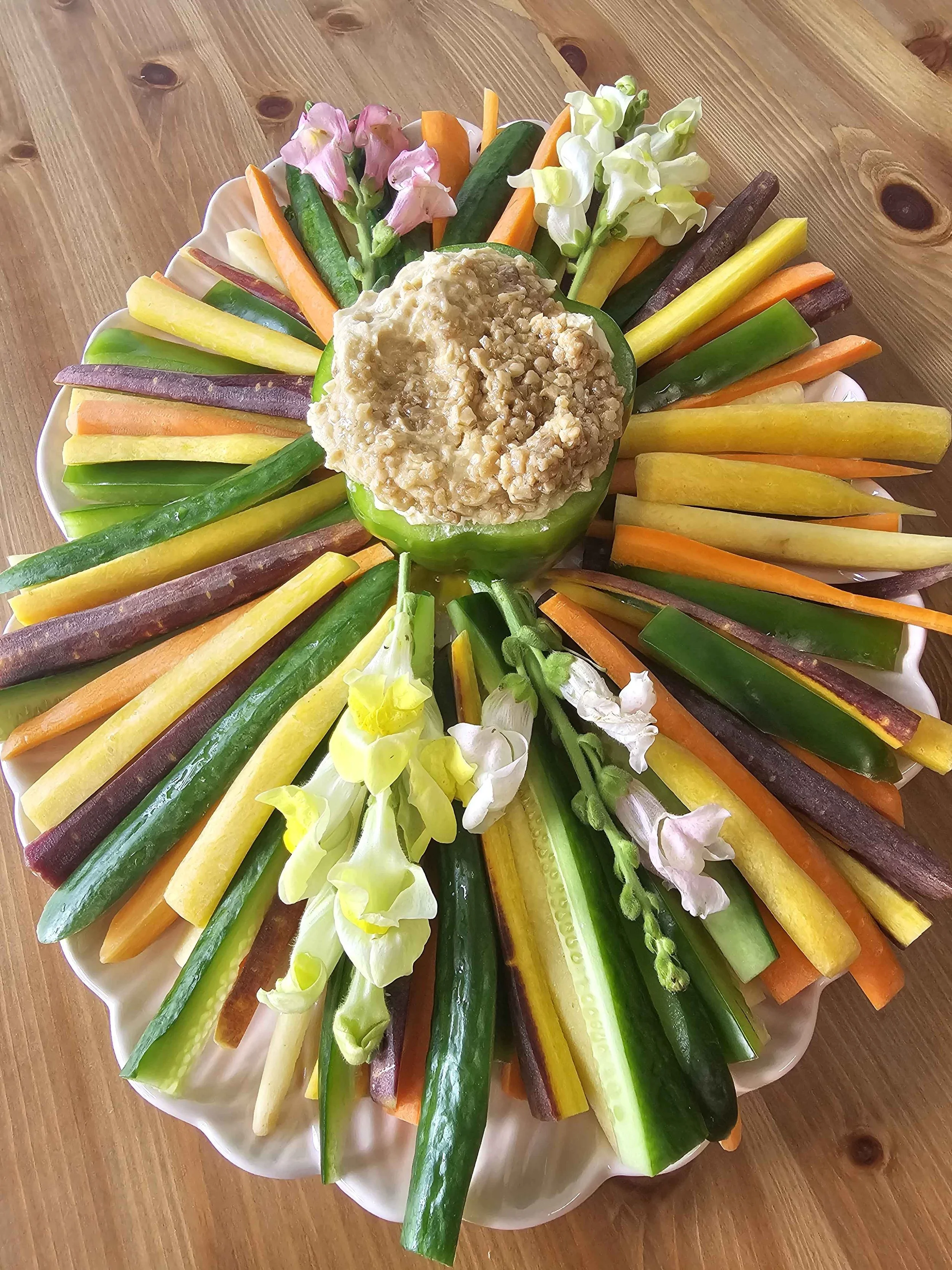 Colorful vegetable platter with sliced cucumbers, carrots, yellow squash, purple sweet potatoes, and green beans arranged in a circle around a dip in a white bowl, garnished with flowers.
