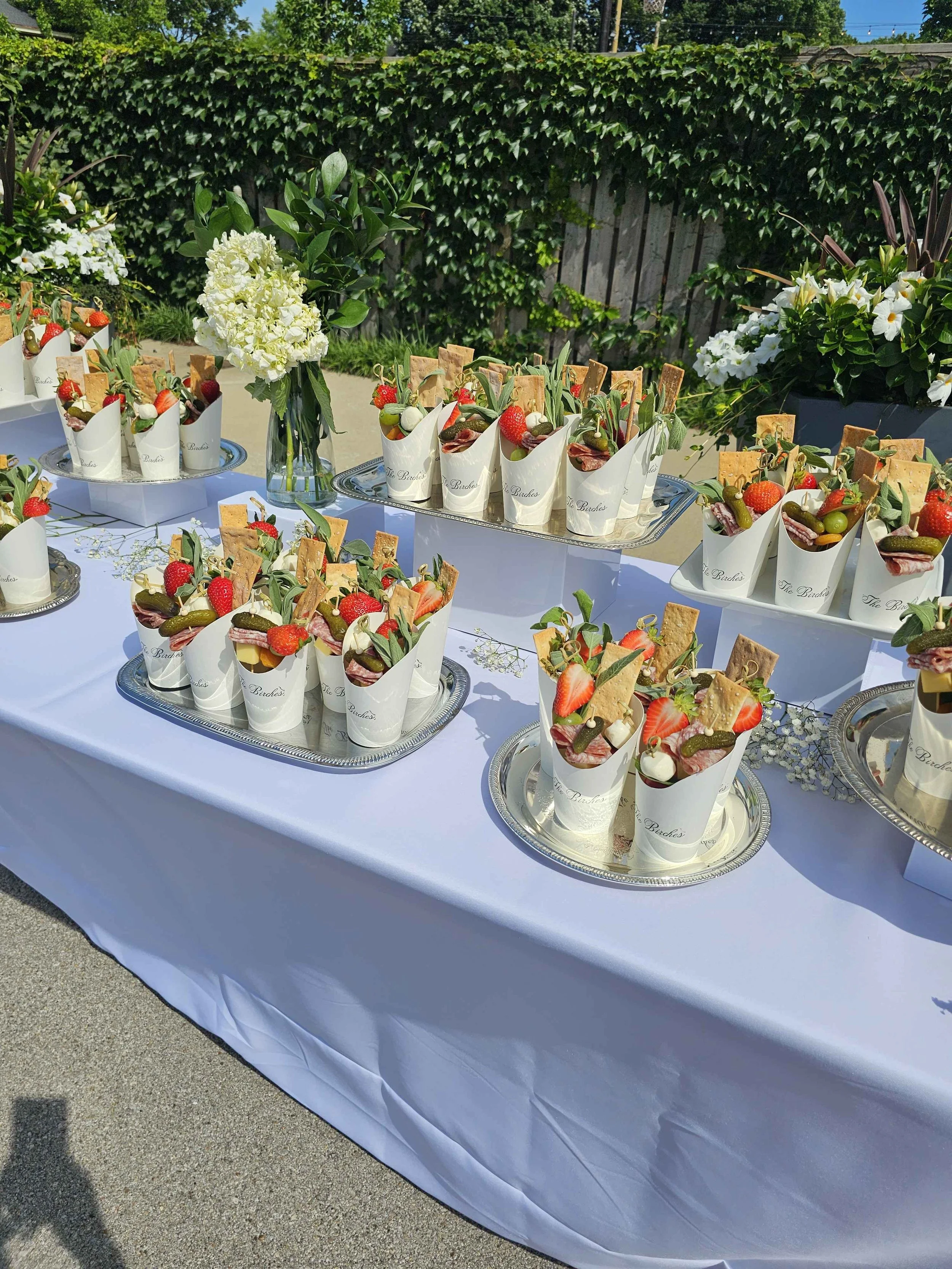 A table covered with a white tablecloth displays small white paper cones filled with strawberries, grapes, prosciutto, and crackers. There are floral arrangements with white hydrangeas and greenery, and the background shows a green hedge and a wooden