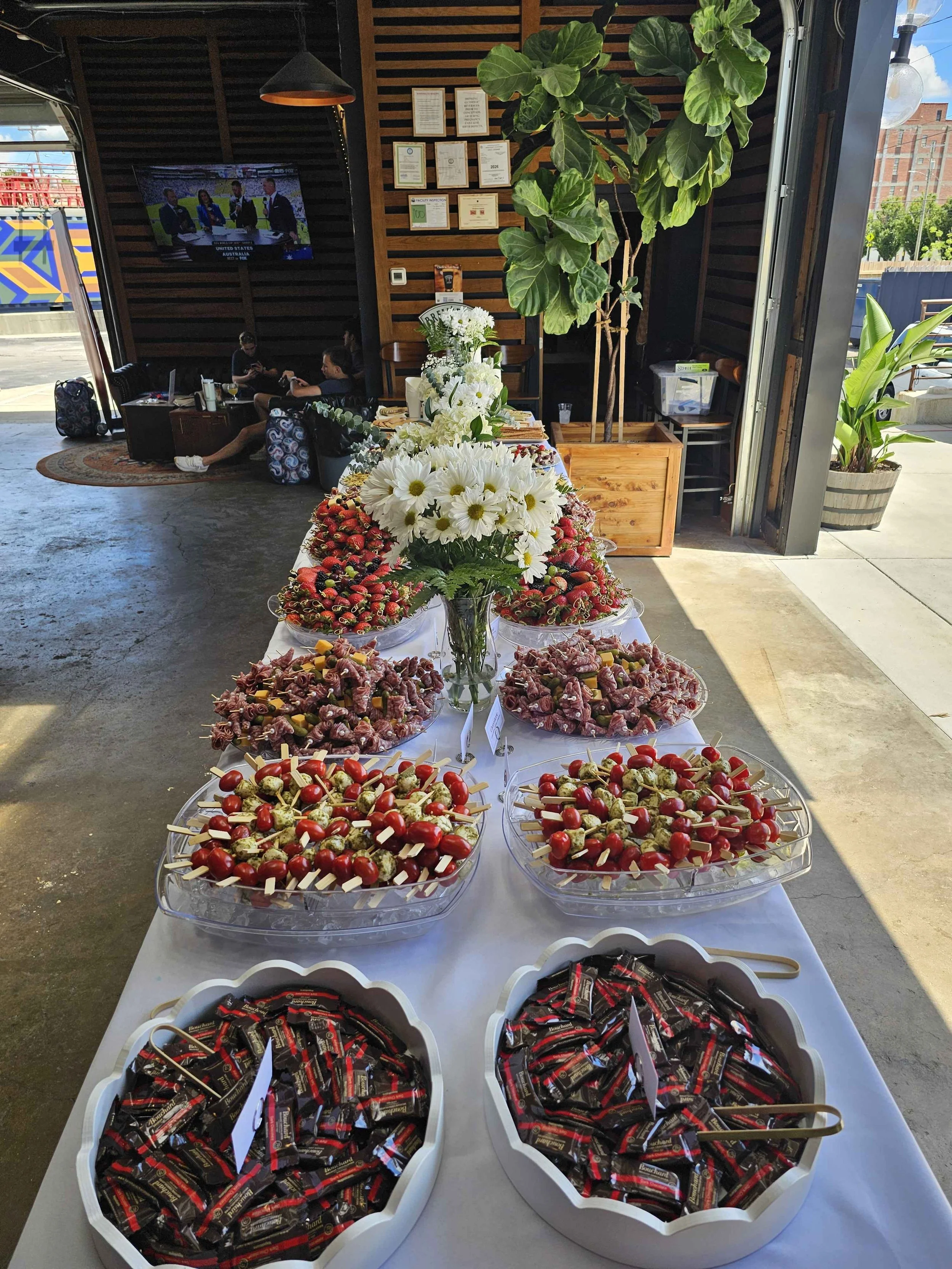 A table displays assorted foods and snacks, including cherry tomatoes, strawberries, cheese cubes, and wrapped chocolates, with bouquets of white daisies and greenery; a living room with two seated people, a TV, framed certificates, and large green p