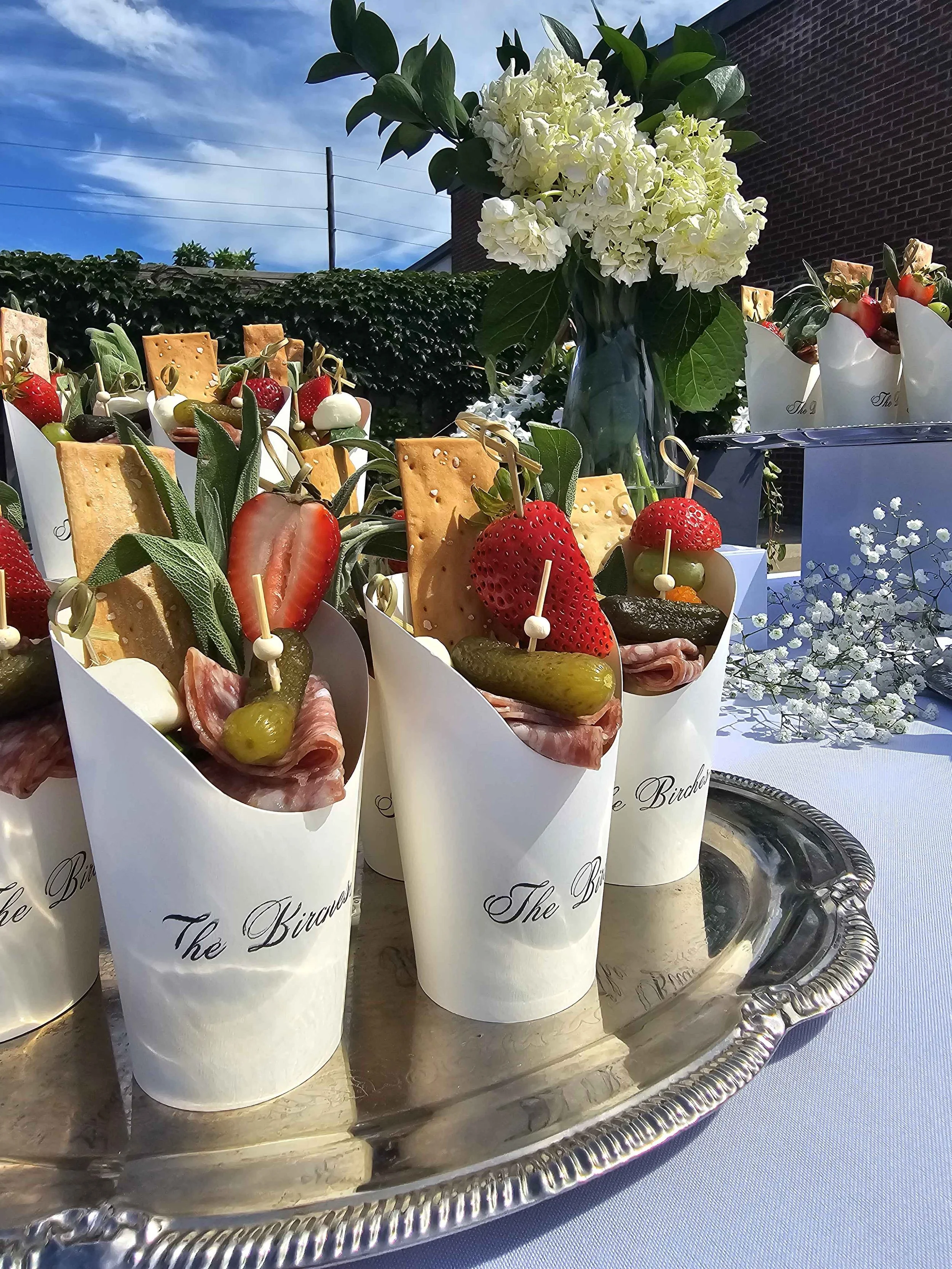 A silver tray holding several charcuterie cups filled with an assortment of foods including strawberries, cheese, olives, and cured meats, with a bouquet of white flowers in the background, outdoors with a blue sky. Made by Rose & Brie Charcuterie.
