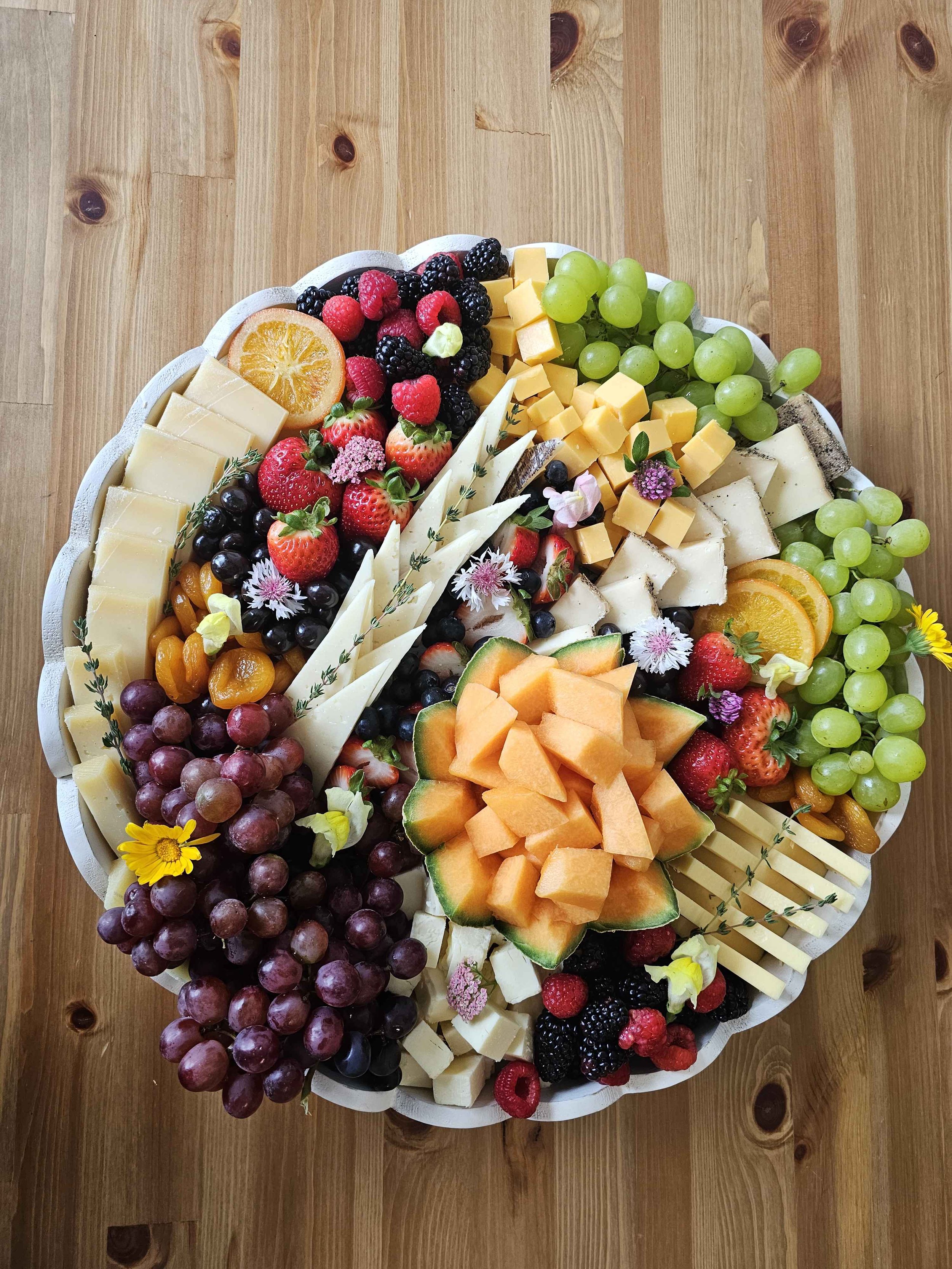 A cheese and fruit platter on a wooden table with green grapes, strawberries, blackberries, raspberries, cantaloupe, orange slices, cheese cubes, and decorative flowers.