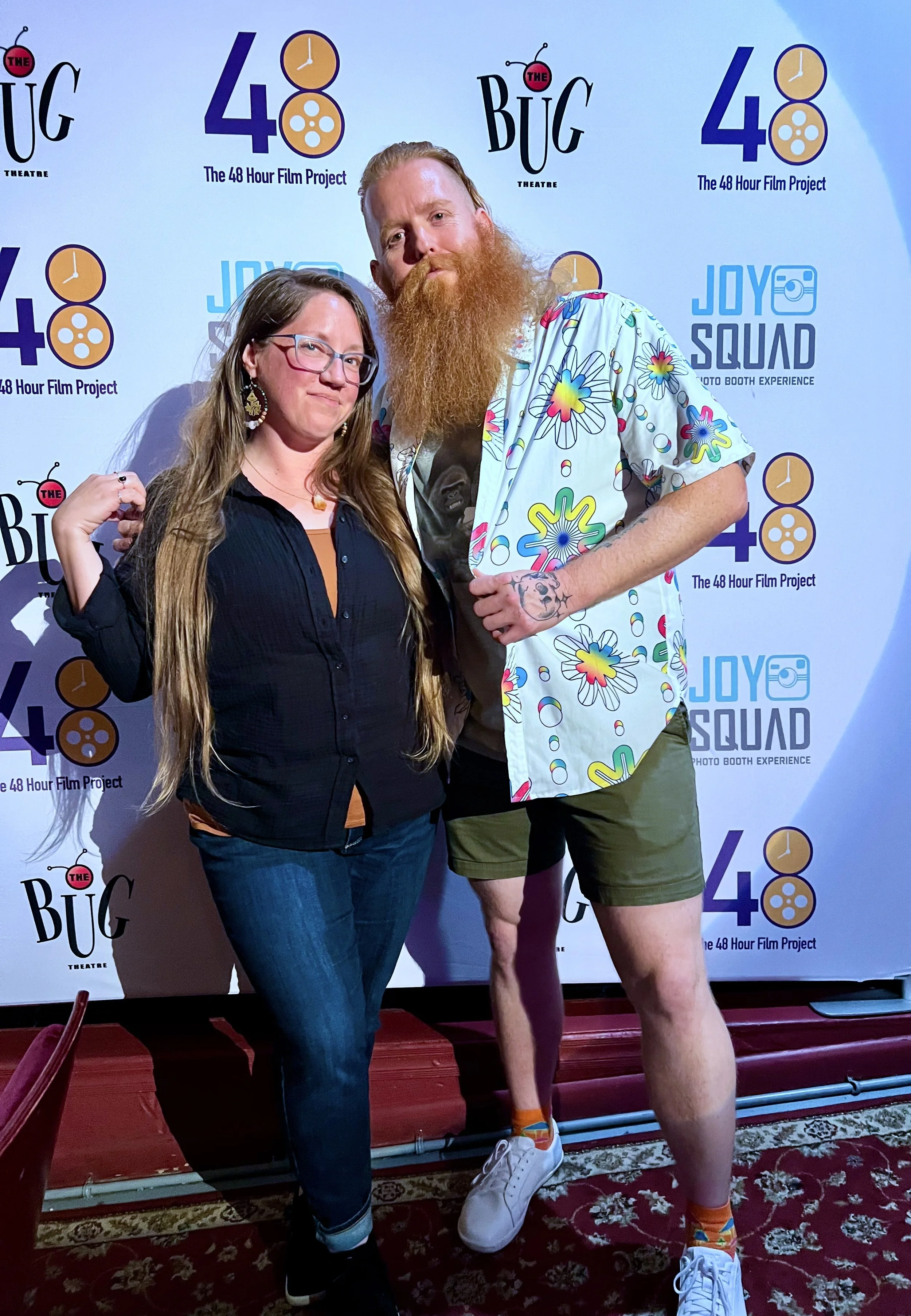 Two people standing in front of a promotional backdrop for the 48 Hour Film Project, with a woman in glasses and a black shirt and a tall man with a long red beard, wearing a colorful floral shirt and shorts.