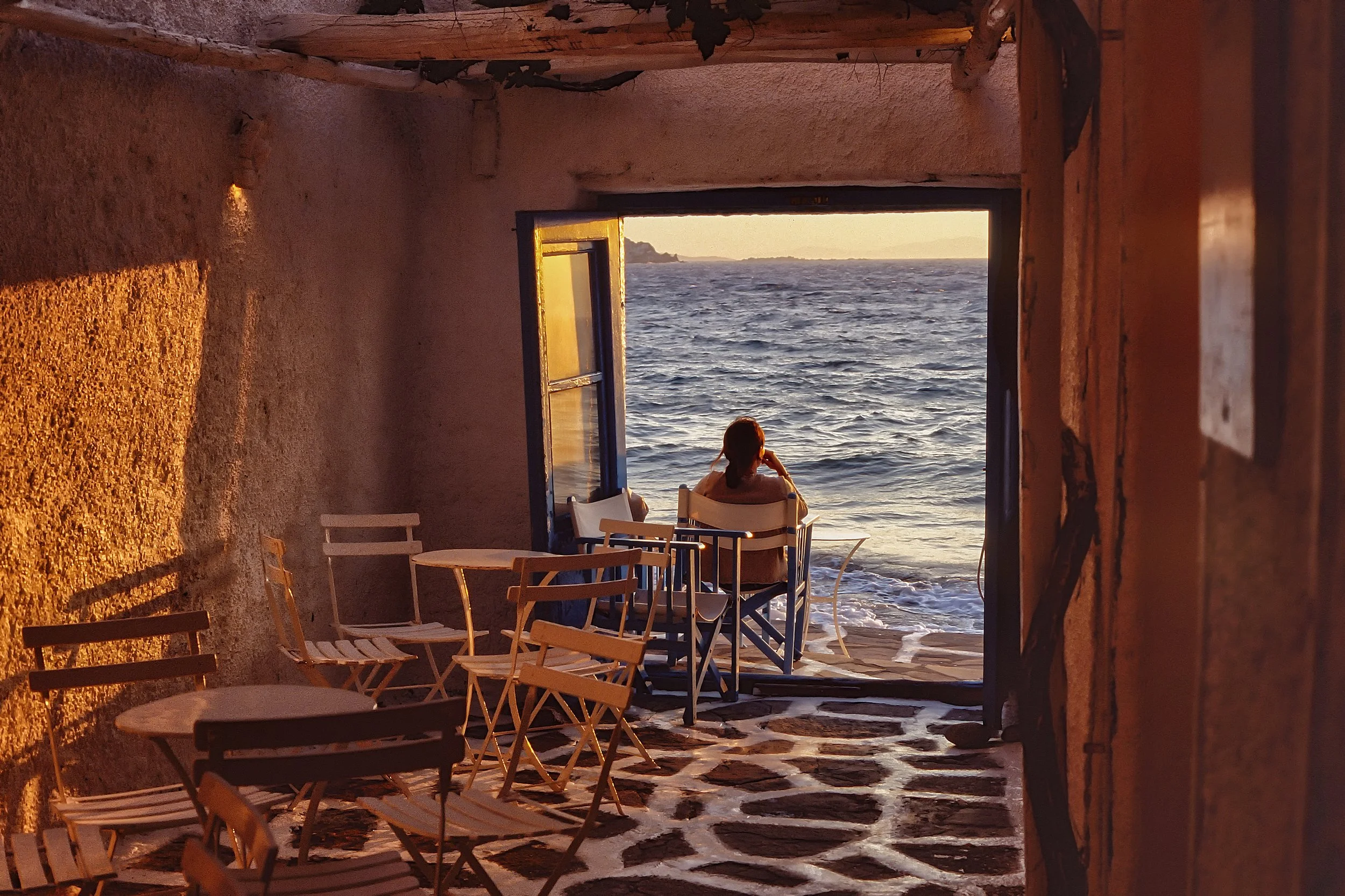 A person sitting on a chair looking out at the ocean through an open doorway in a rustic room with stone floor and wooden ceiling at sunset.