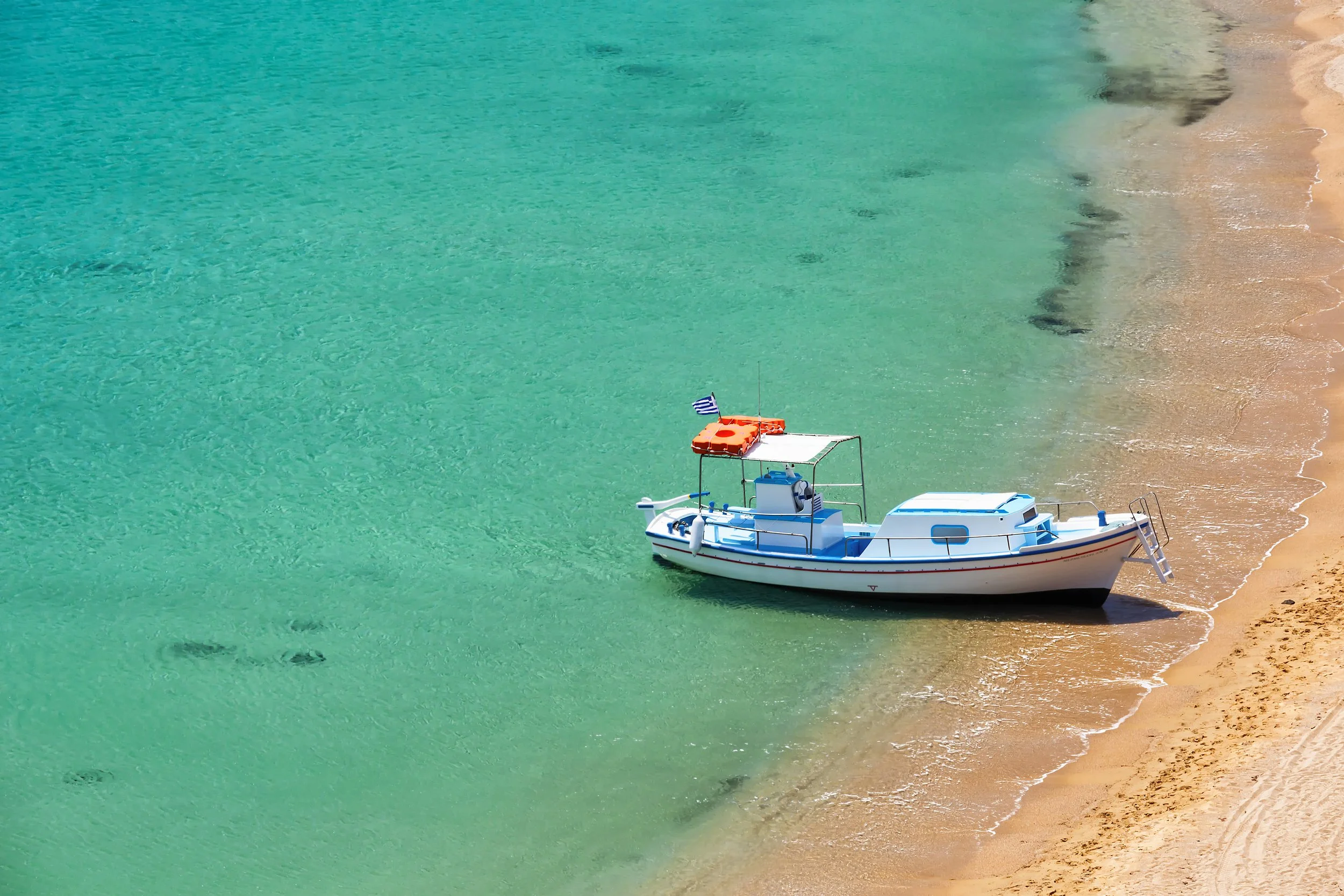A small white boat floating near the sandy shoreline of a calm, turquoise sea with footprints in the sand nearby.