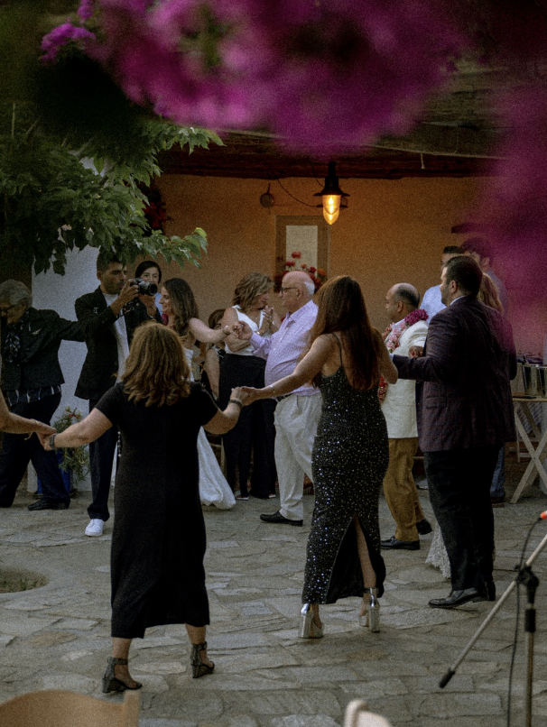 People dancing and celebrating at a social gathering or party outdoors in the evening, with colorful flowers and string lights in the background.