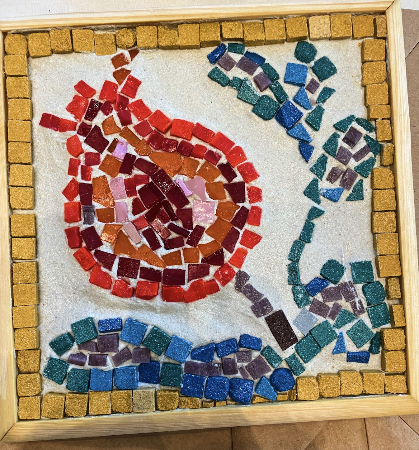 Mosaic artwork of a red and pink rose with blue and teal butterflies, bordered by small yellow tiles, on a white sandy background.