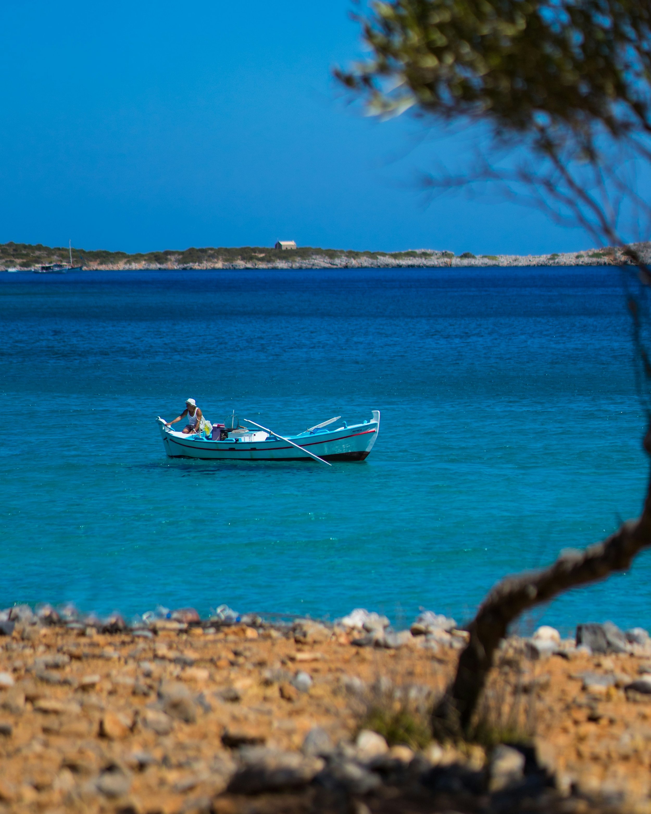 A person in a small white boat on a calm, blue body of water, with a rocky shoreline and a distant landmass with a small building in the background, and some trees in the foreground.