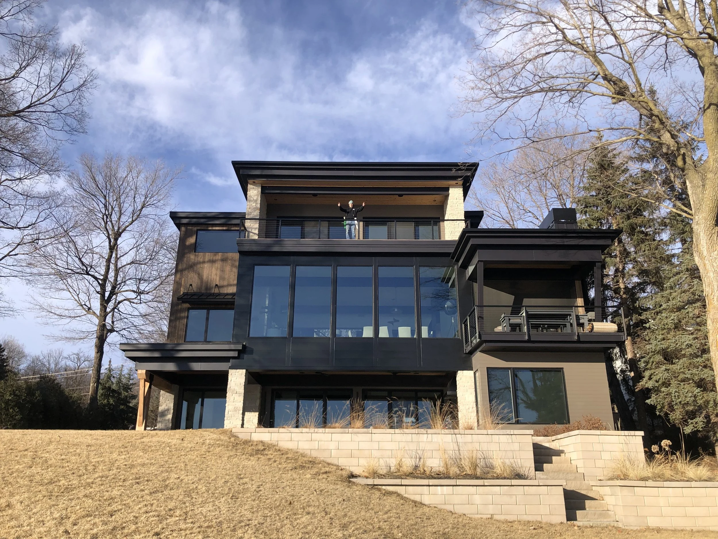 Modern three-story house with large windows, balcony, and surrounding trees