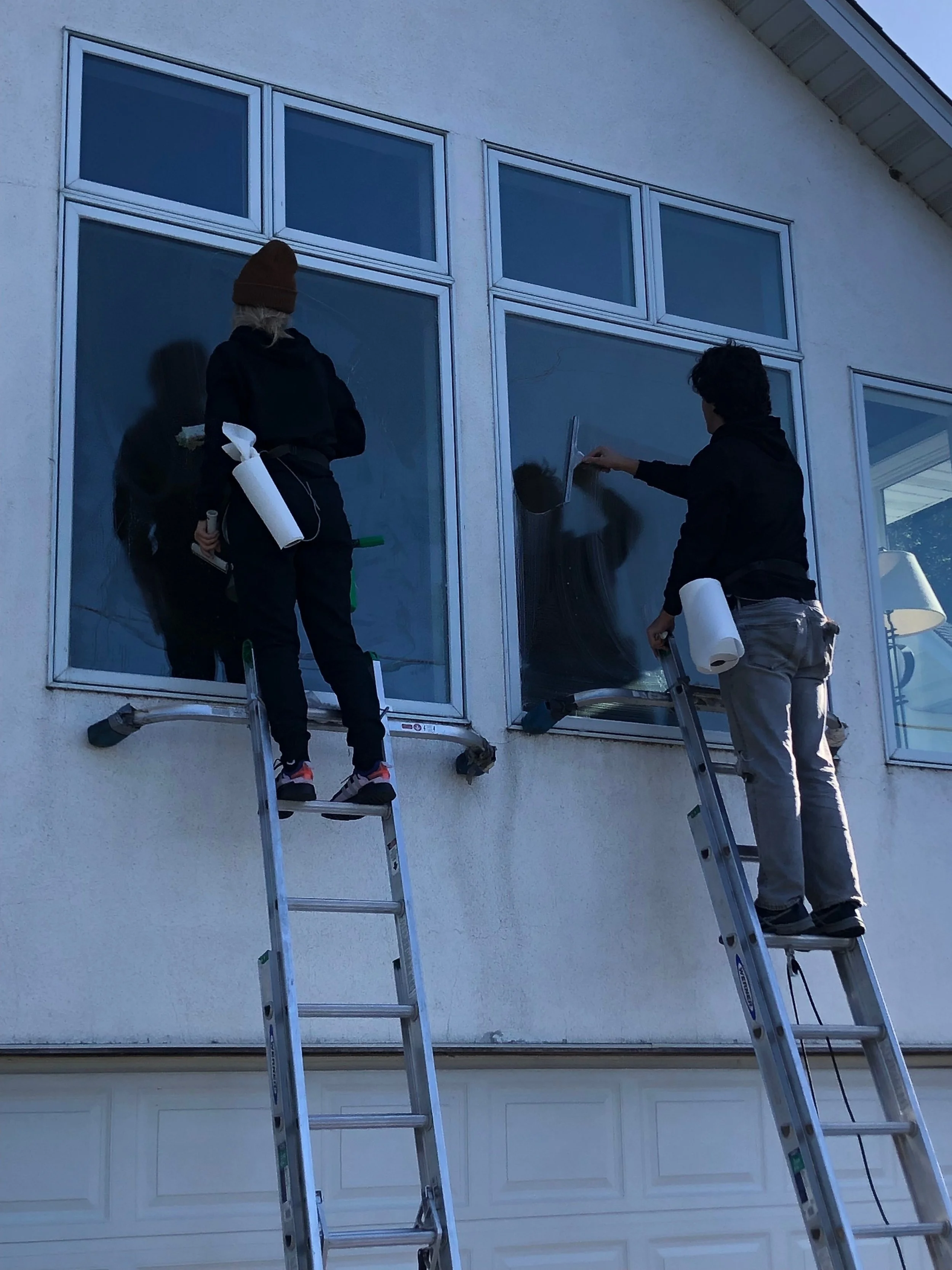 Two people on ladders cleaning large windows of a house, holding cleaning supplies.