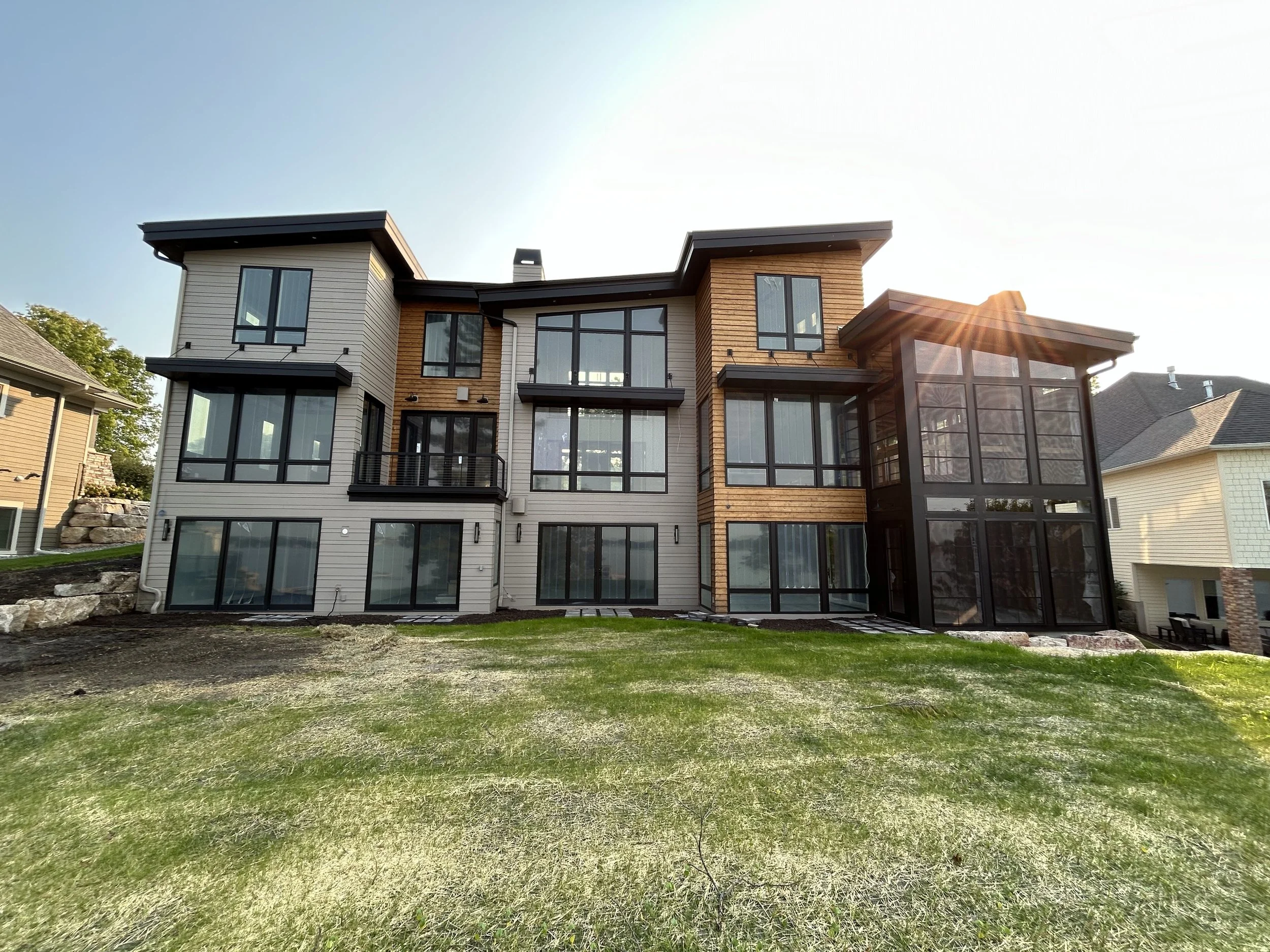 Modern three-story house with large windows, wooden accents, and a manicured lawn on a sunny day.