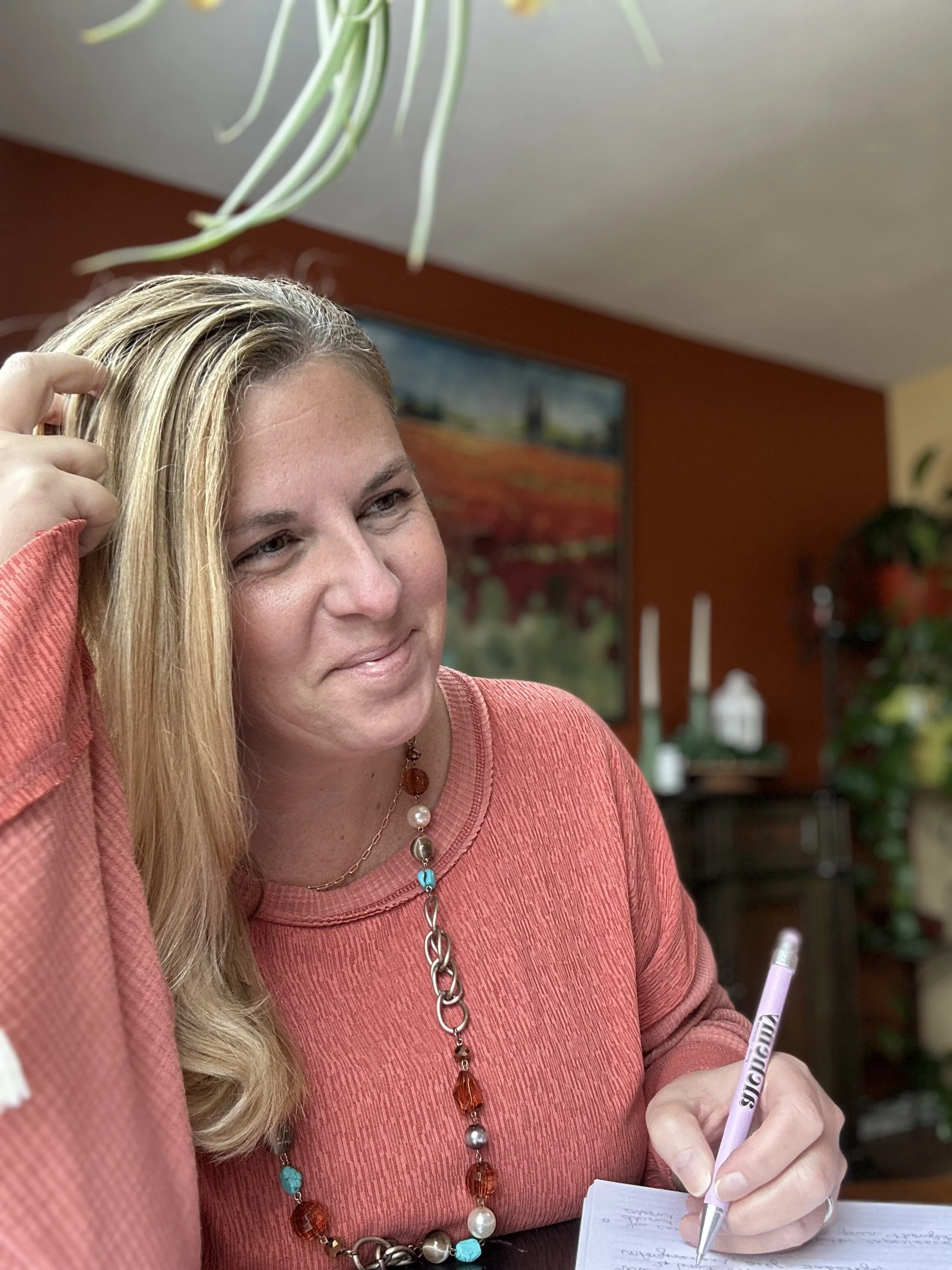 A woman with blonde hair wearing a coral-colored top, sitting at a table, writing on a notepad with a purple pen.