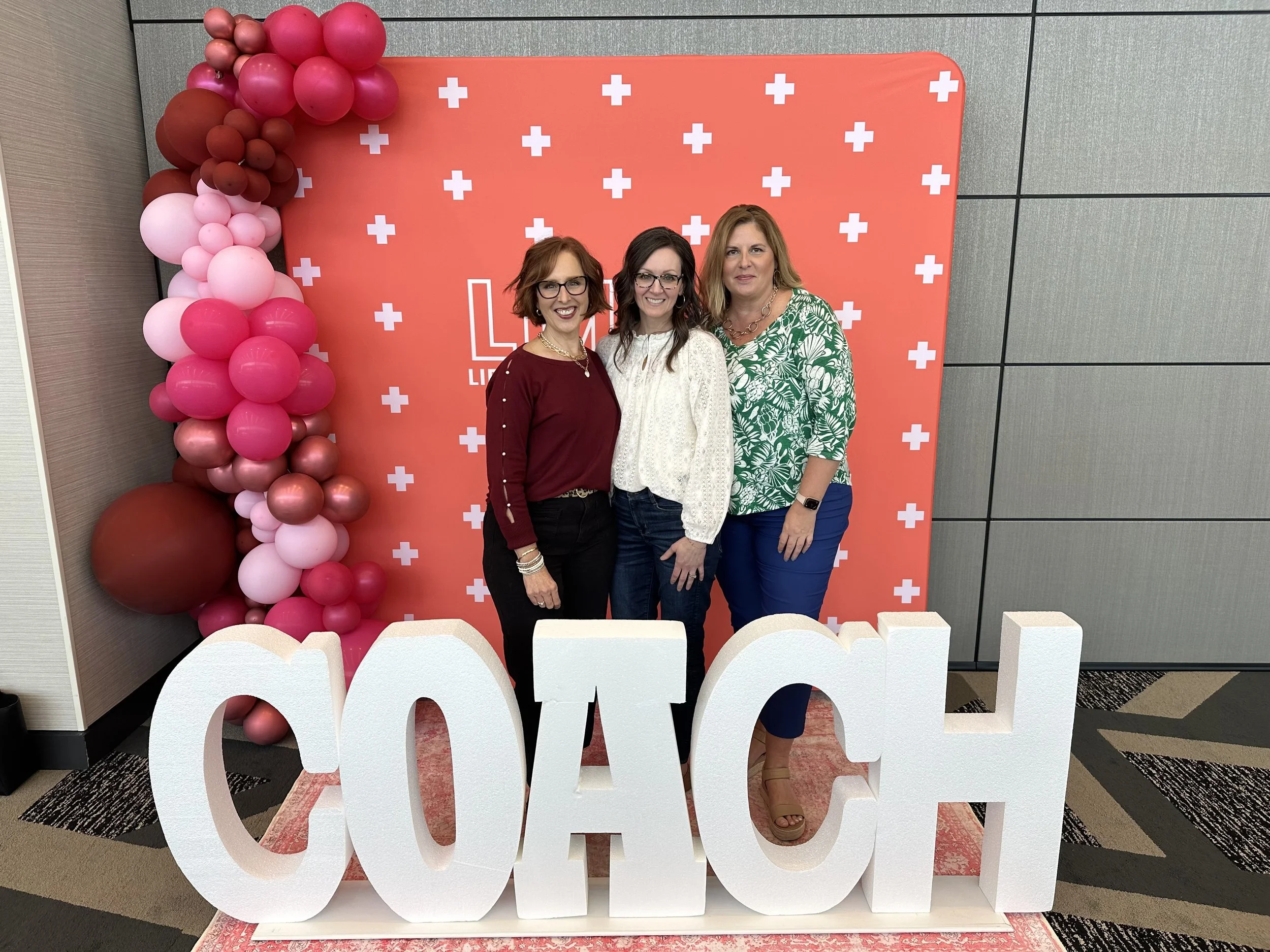 Three women standing behind large white letters spelling out 'COACH', with a pink backdrop decorated with white plus signs and a balloon arrangement on the left side.