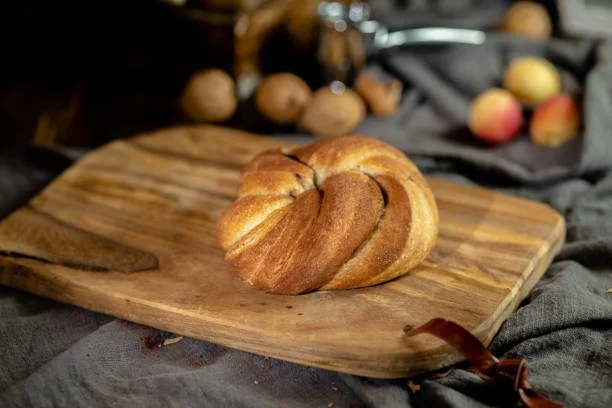 A croissant on a wooden cutting board with assorted nuts and peaches in the background.