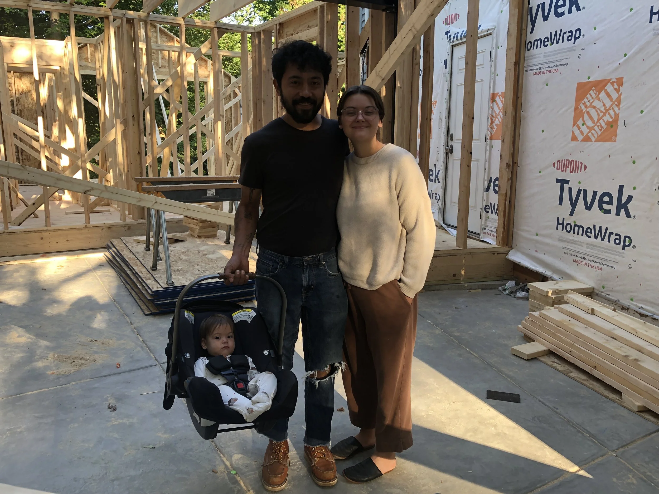 A family consisting of an adult holding a baby in a car seat and another adult standing next to them inside a house under construction, with visible framing and Tyvek wrap.