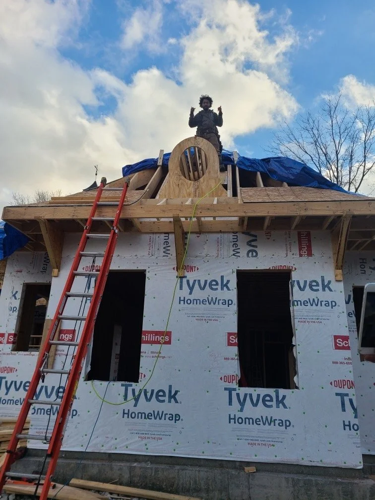 Construction site with a partially built house using Tyvek HomeWrap and blue tarps. A person stands on the roof next to a circular window opening. A red ladder leans against the structure.