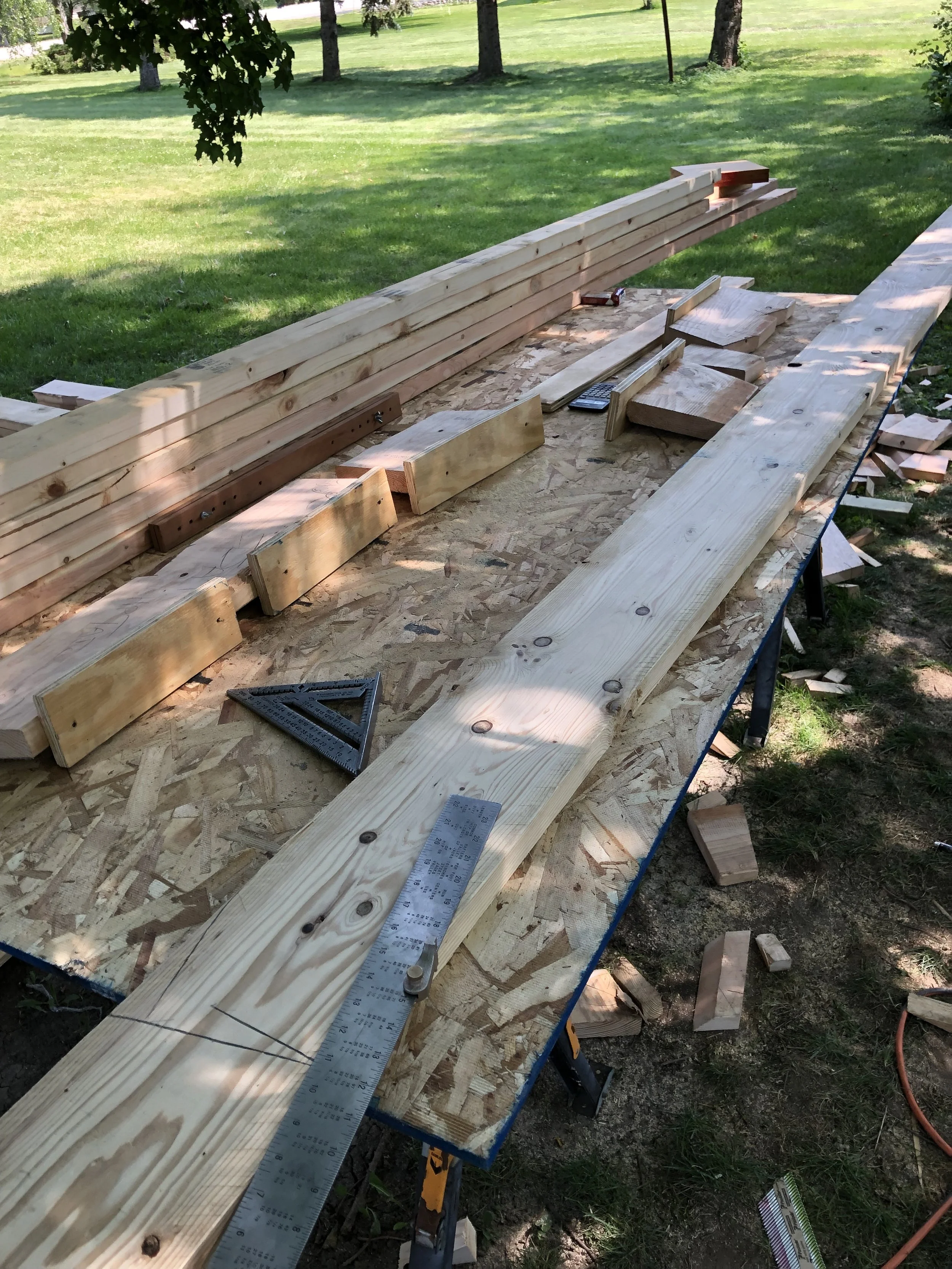 Outdoor woodworking setup with lumber, a measuring square, and a ruler on a sawhorse table.