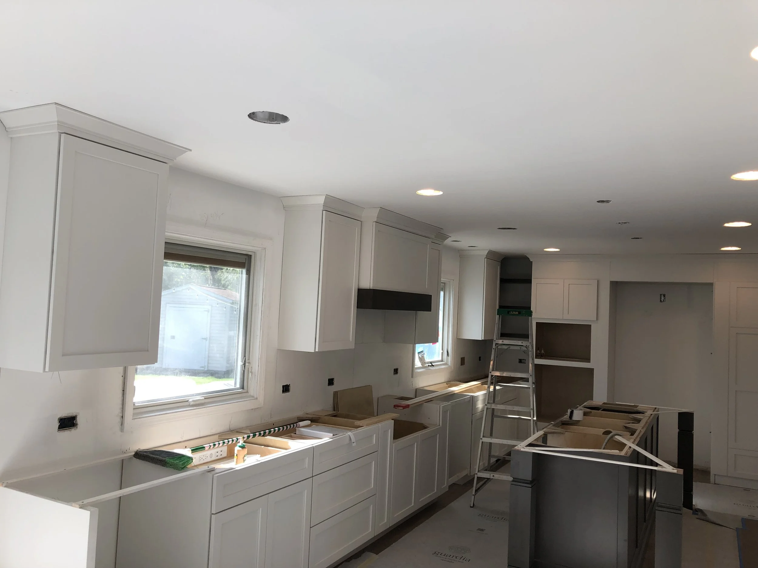 A partially renovated modern kitchen with white cabinetry, a window, and recessed lighting. A ladder and construction materials are present, indicating ongoing work.