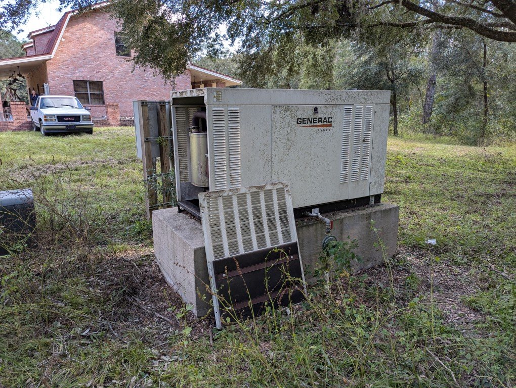 An outdoor scene with a Generac generator on a concrete pad in a grassy yard near a house, with a tree overhead and a car parked on the driveway.